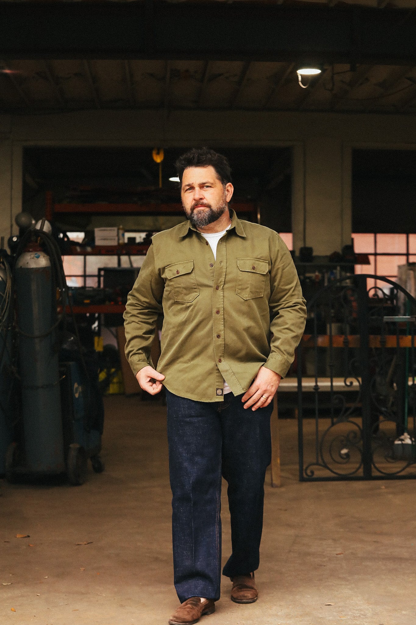 Man wearing a green jacket and blue jeans standing in an indoor setting with equipment in the background.