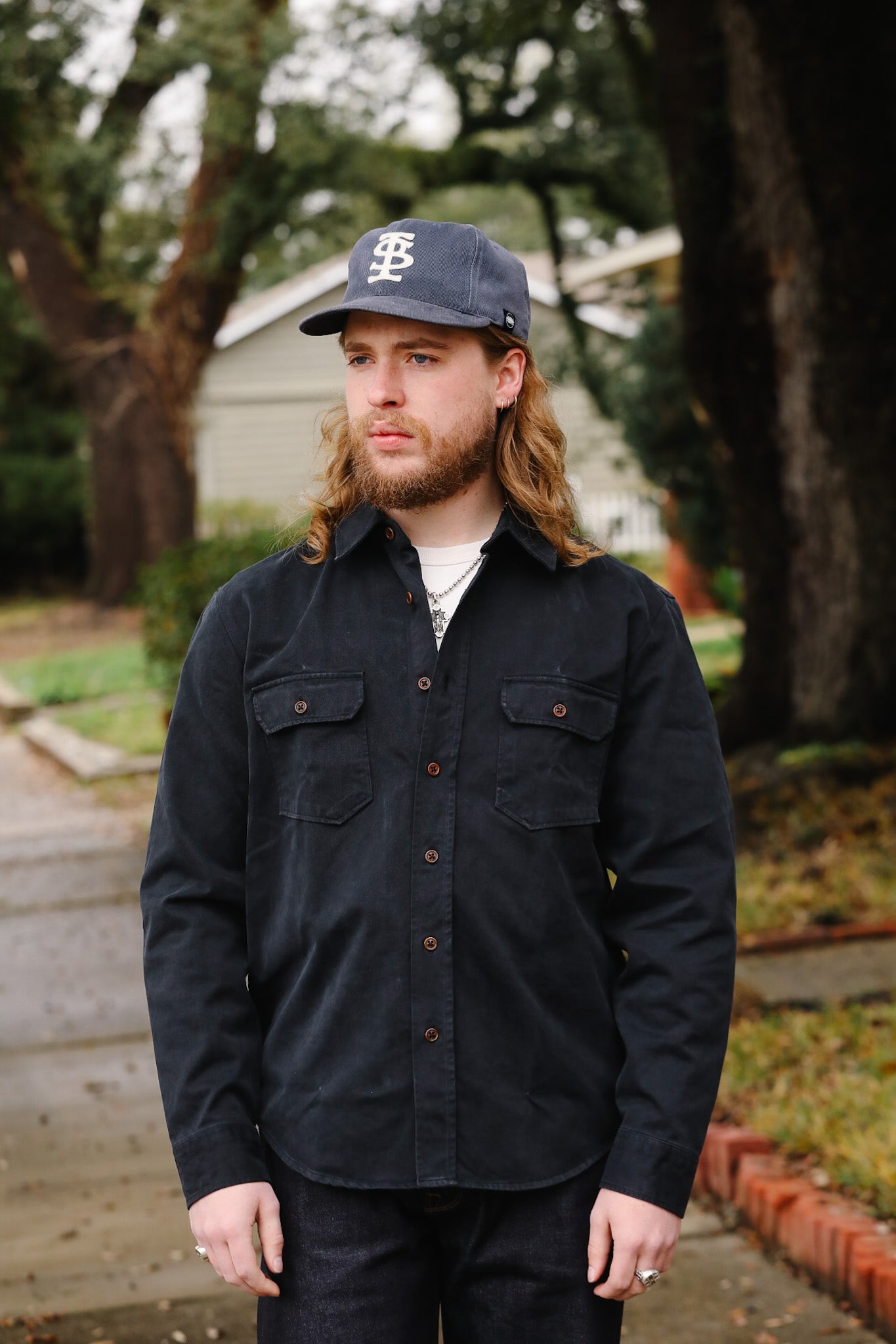 Man wearing a black jacket and cap outdoors with trees and a building in the background