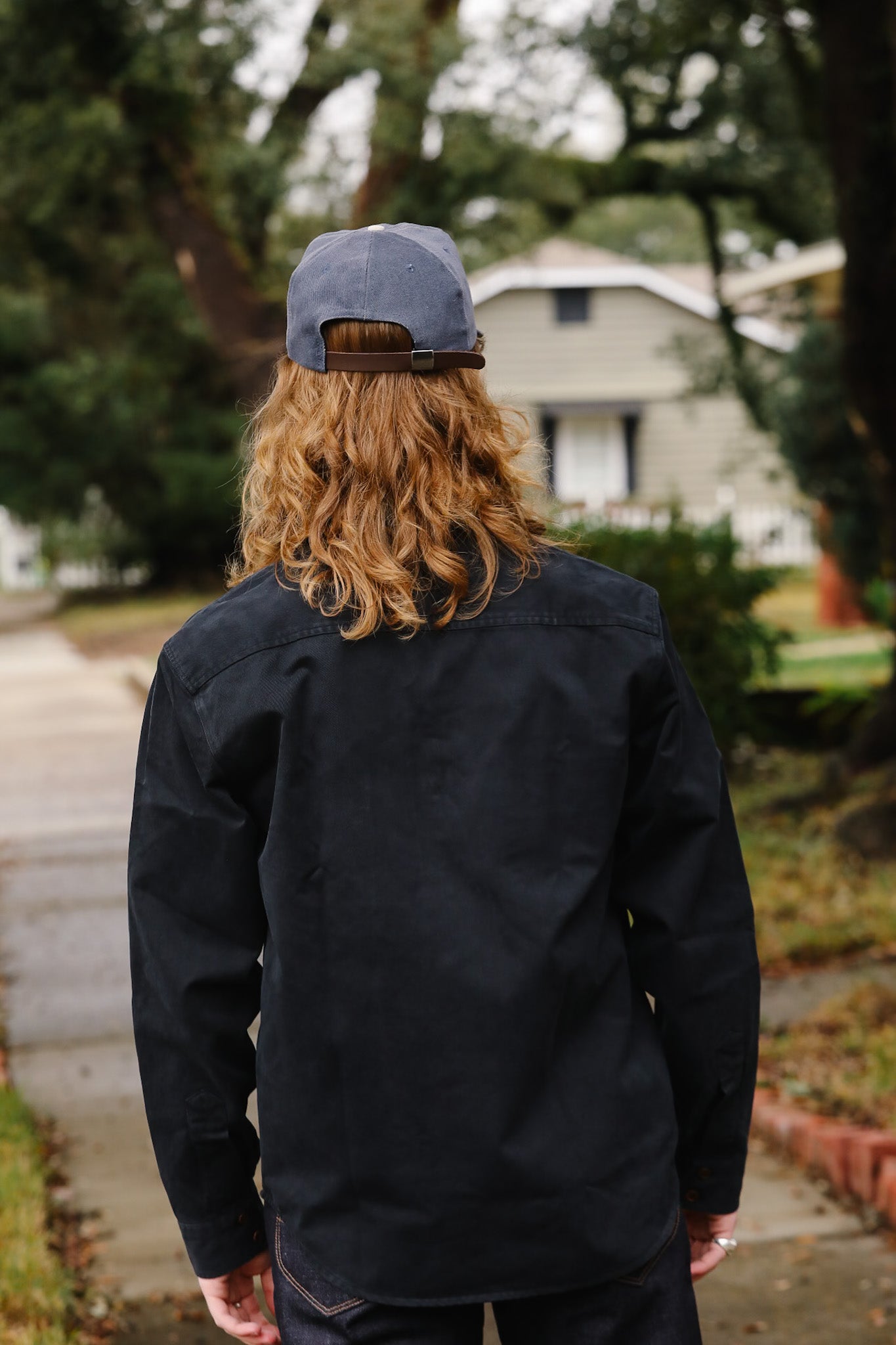 Person wearing a black jacket and blue cap walking away from the camera with a residential area in the background.