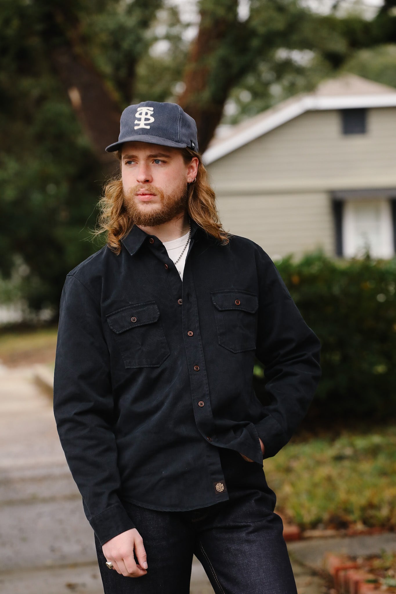 Man wearing a black jacket and cap standing outdoors with a house and trees in the background