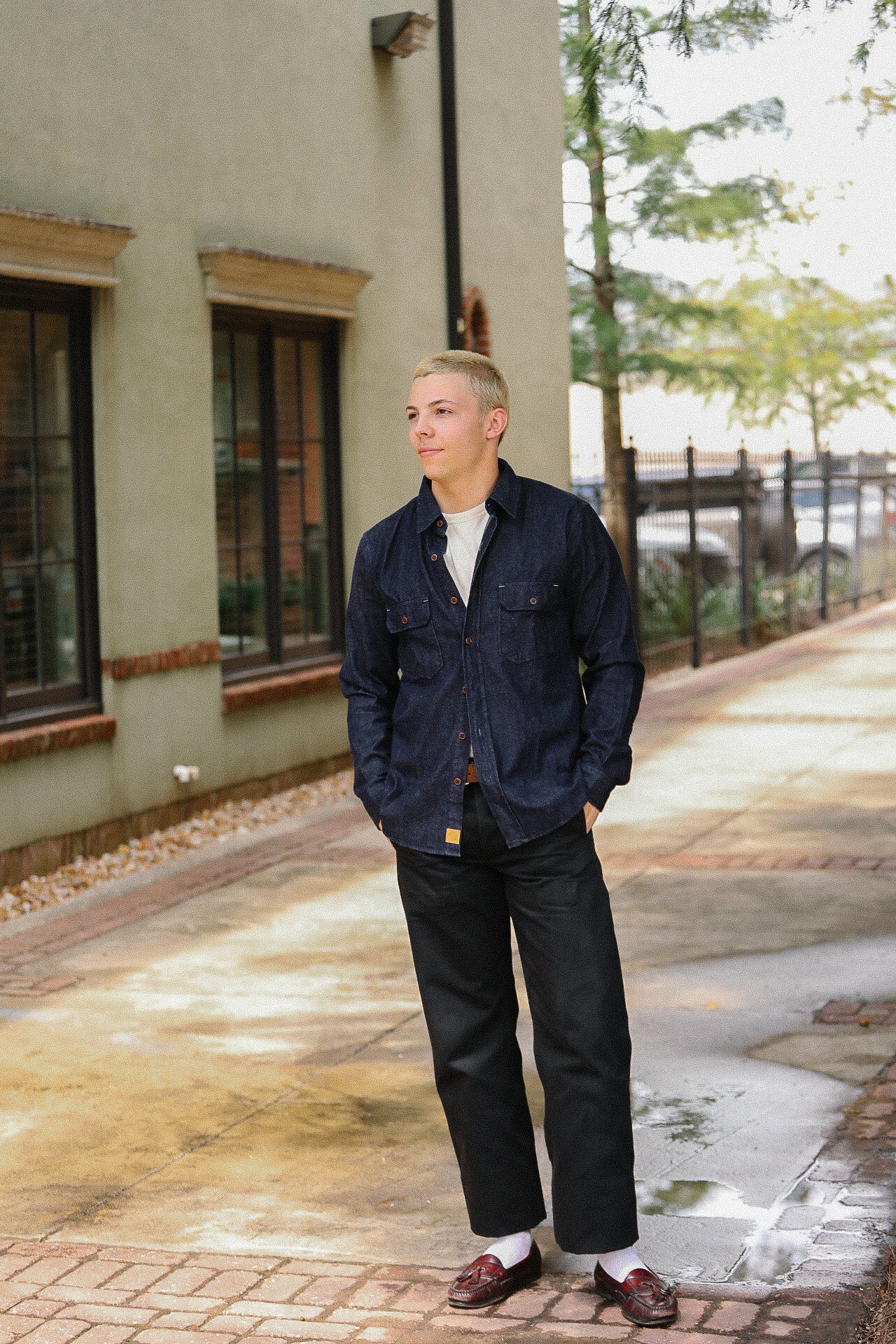 Young man wearing denim shirt, black pants, white socks, and brown leather loafers standing outdoors on brick pathway
