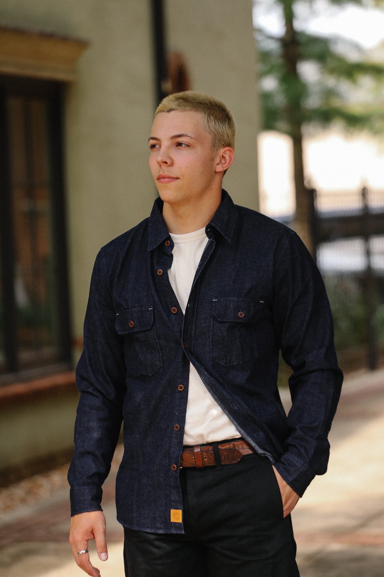 Man wearing dark denim shirt over white tee with brown belt standing outdoors