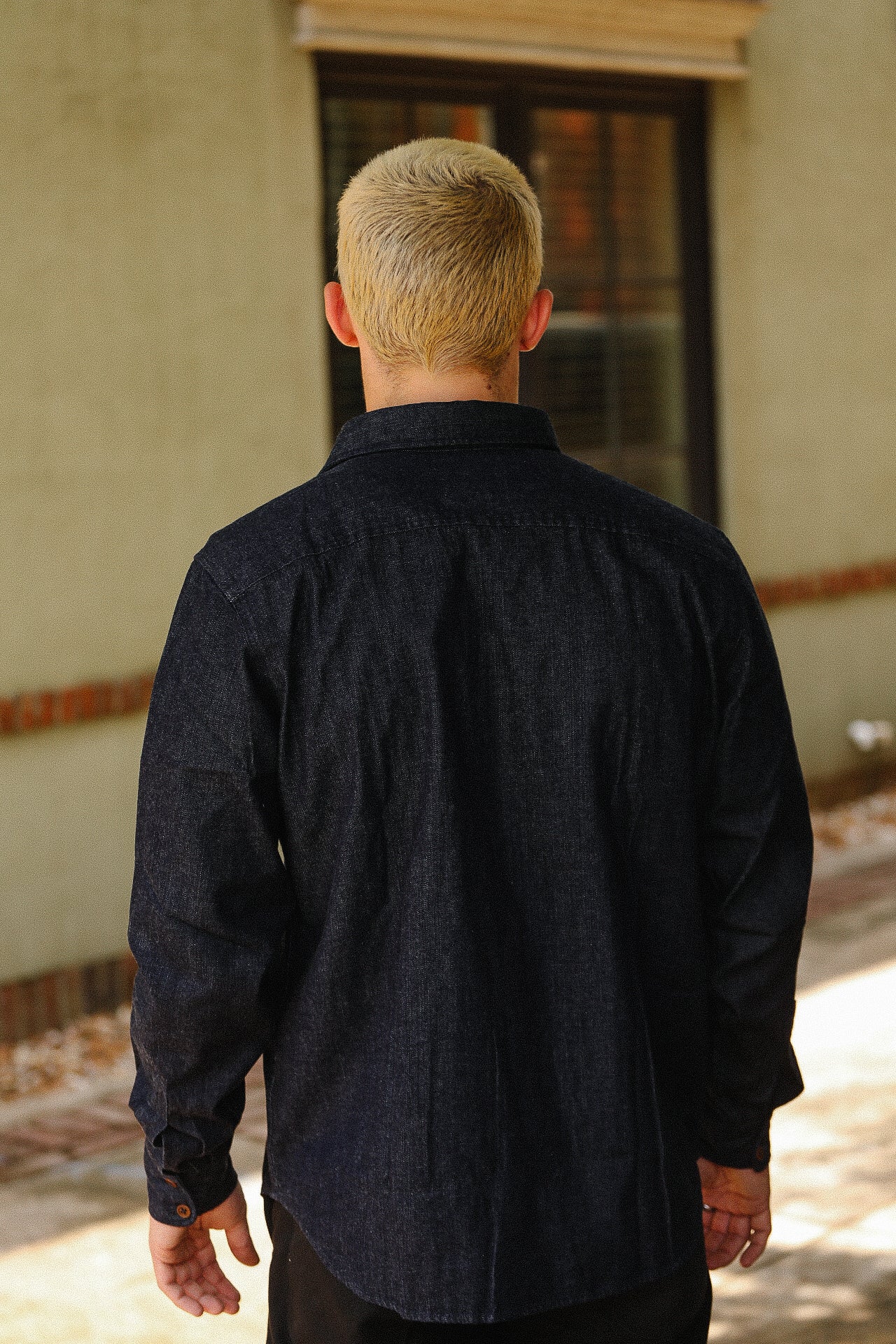 Man with blonde hair wearing a dark denim shirt standing outdoors near a window