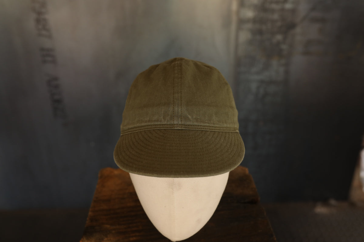 An olive green cotton baseball cap displayed on a white mannequin head resting on a wooden surface, with a dark, textured studio background.