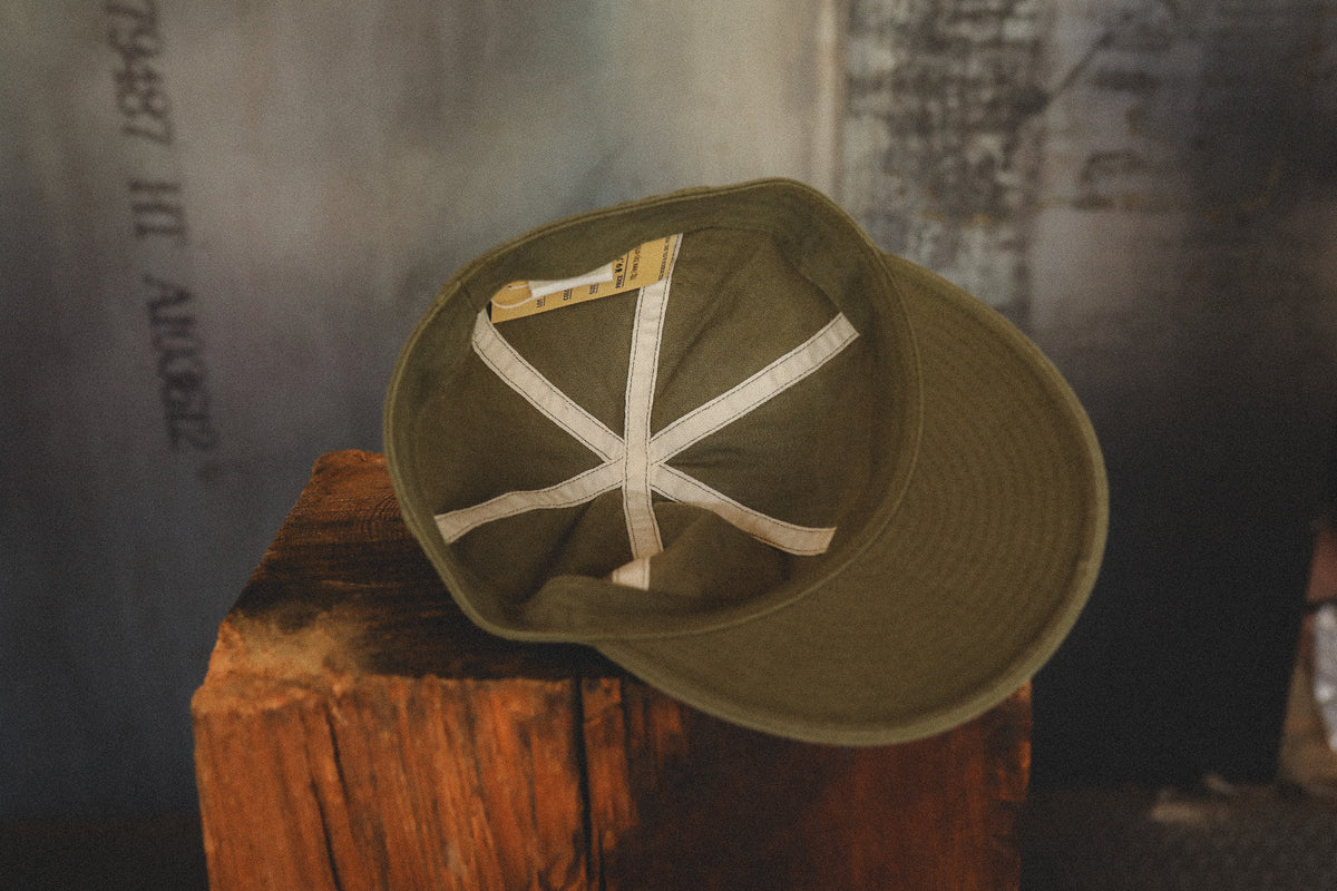 An olive green baseball cap with a white cross brim, resting on a rustic wooden block against a dark, textured background.