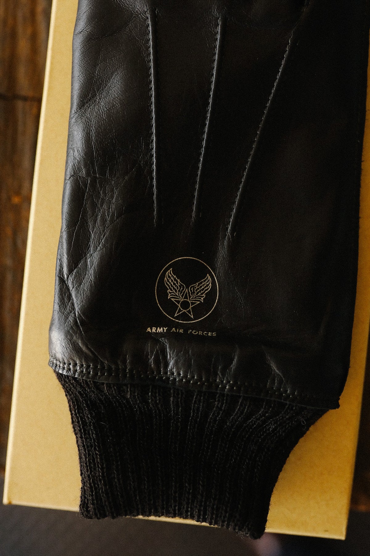 Close-up of a black leather jacket sleeve with a ribbed knit cuff, featuring a gold embroidered Army Air Forces emblem and text on the upper cuff.