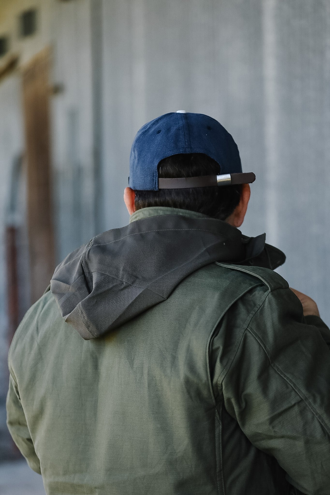 Person wearing a blue cap and green jacket standing against a corrugated metal wall.
