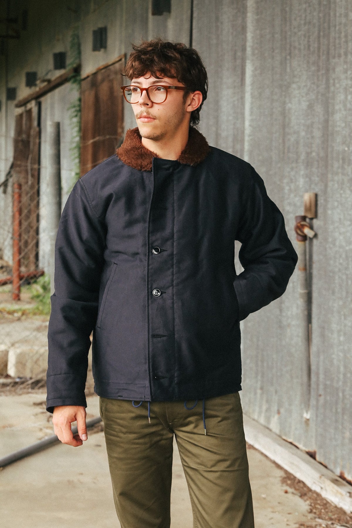 A man wearing a dark navy blue waxed cotton jacket with a brown faux-fur collar, paired with olive green pants, standing in front of a weathered gray metal wall.