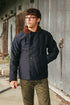 A man wearing a dark navy blue waxed cotton jacket with a brown faux-fur collar, paired with olive green pants, standing in front of a weathered gray metal wall.