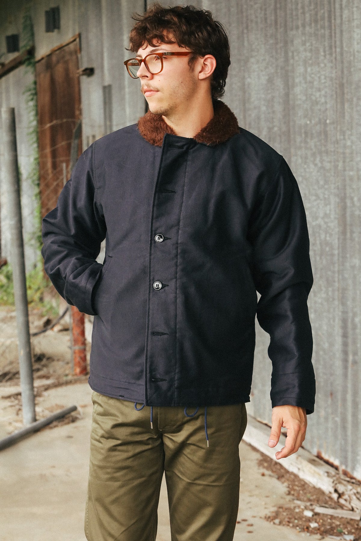 A man wearing a dark navy blue waxed cotton jacket with a brown faux-fur collar, paired with olive green pants, standing in front of a rustic corrugated metal wall.