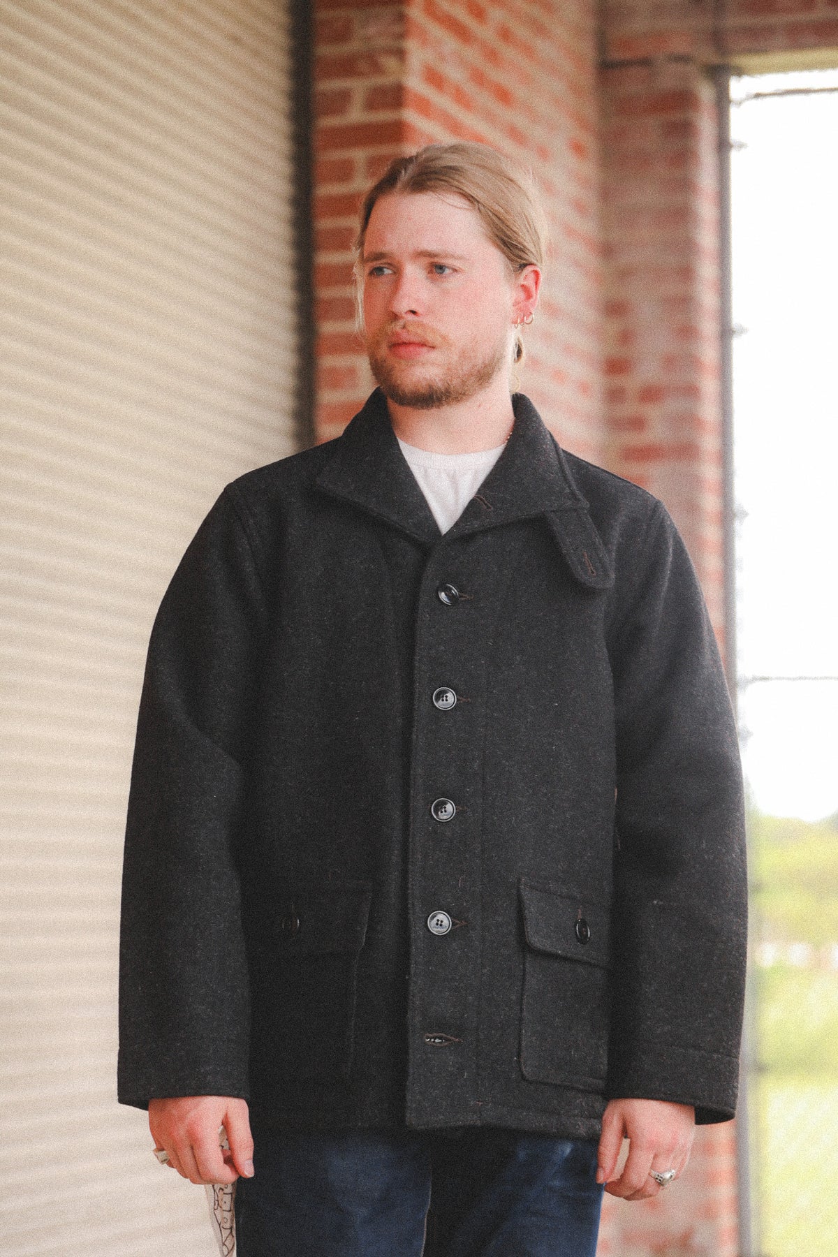 A person  wearing a dark charcoal gray wool-blend coat, standing in front of a red brick wall and a metal roll-up door.
