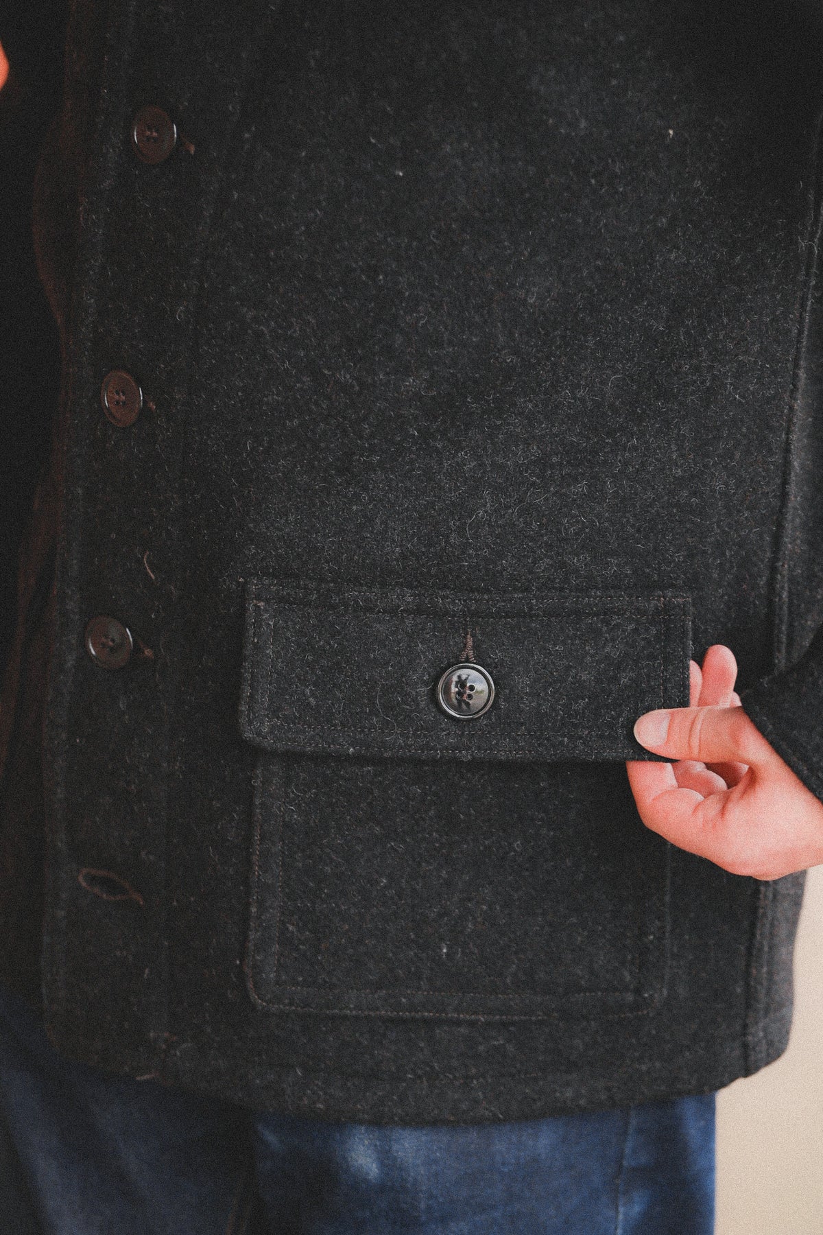 A person  wearing a dark charcoal gray wool-blend coat, standing in front of a red brick wall and a metal roll-up door.