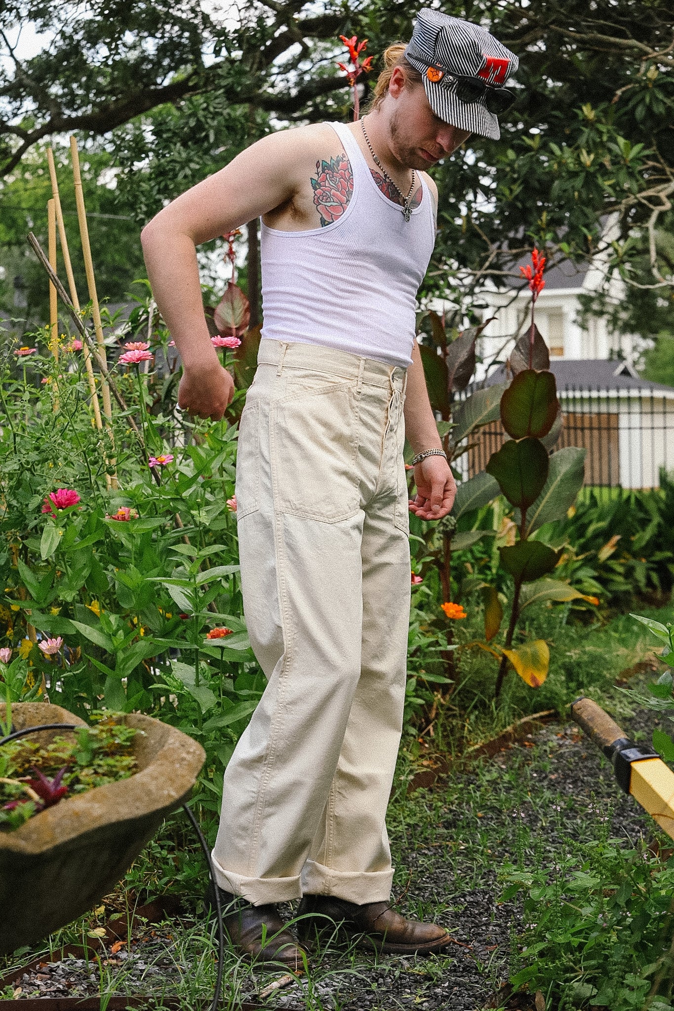 Man in a garden wearing a white tank top and beige pants, surrounded by plants and flowers.