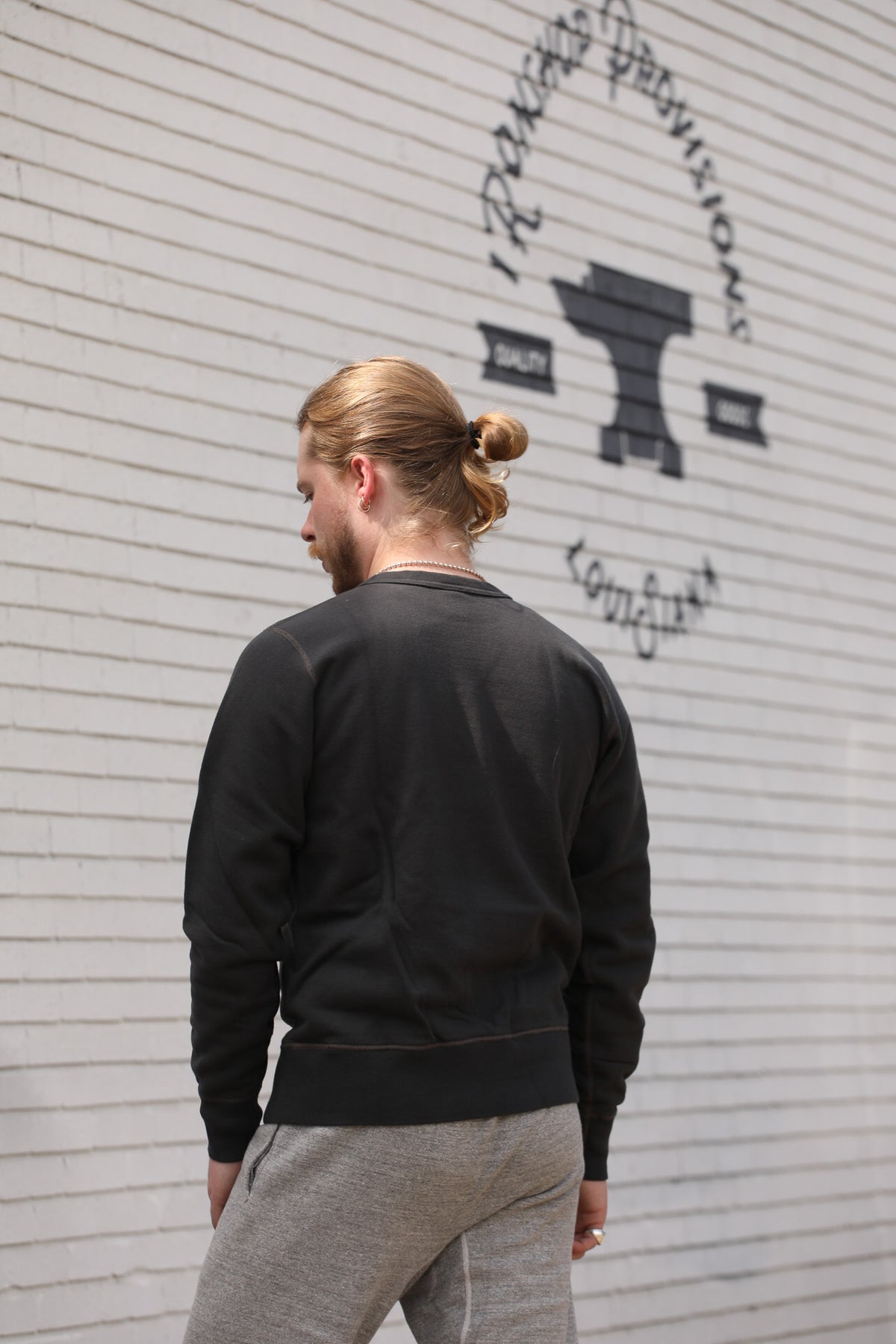 A man with blonde hair in a bun is seen from behind, wearing a black crewneck sweatshirt and gray sweatpants, standing in front of a white brick wall with a black logo.