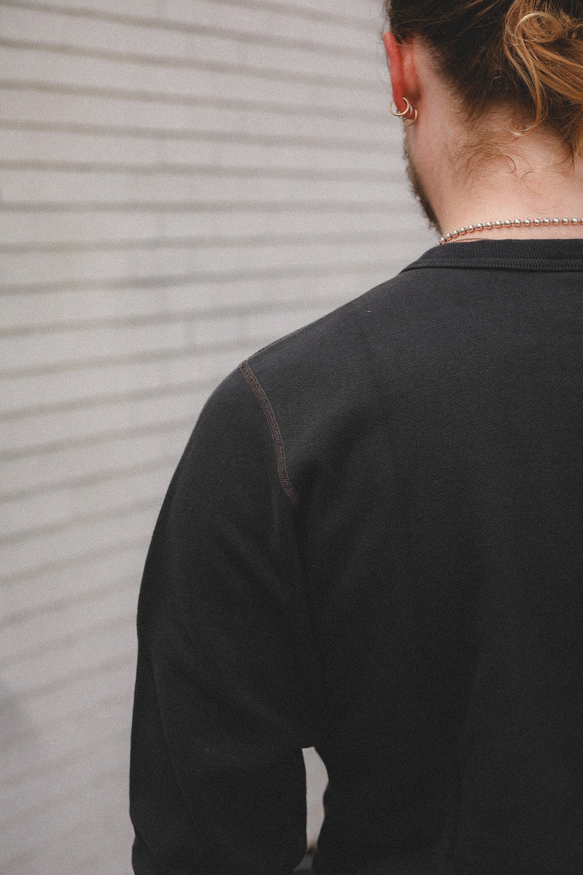 A person shown from behind wearing a black long-sleeve crewneck sweatshirt, standing against a light gray textured wall.