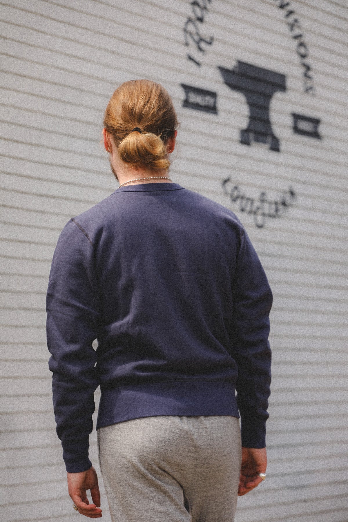A person viewed from behind wearing a navy blue crewneck sweatshirt and light gray sweatpants, standing in front of a white corrugated metal wall with a black graphic logo.