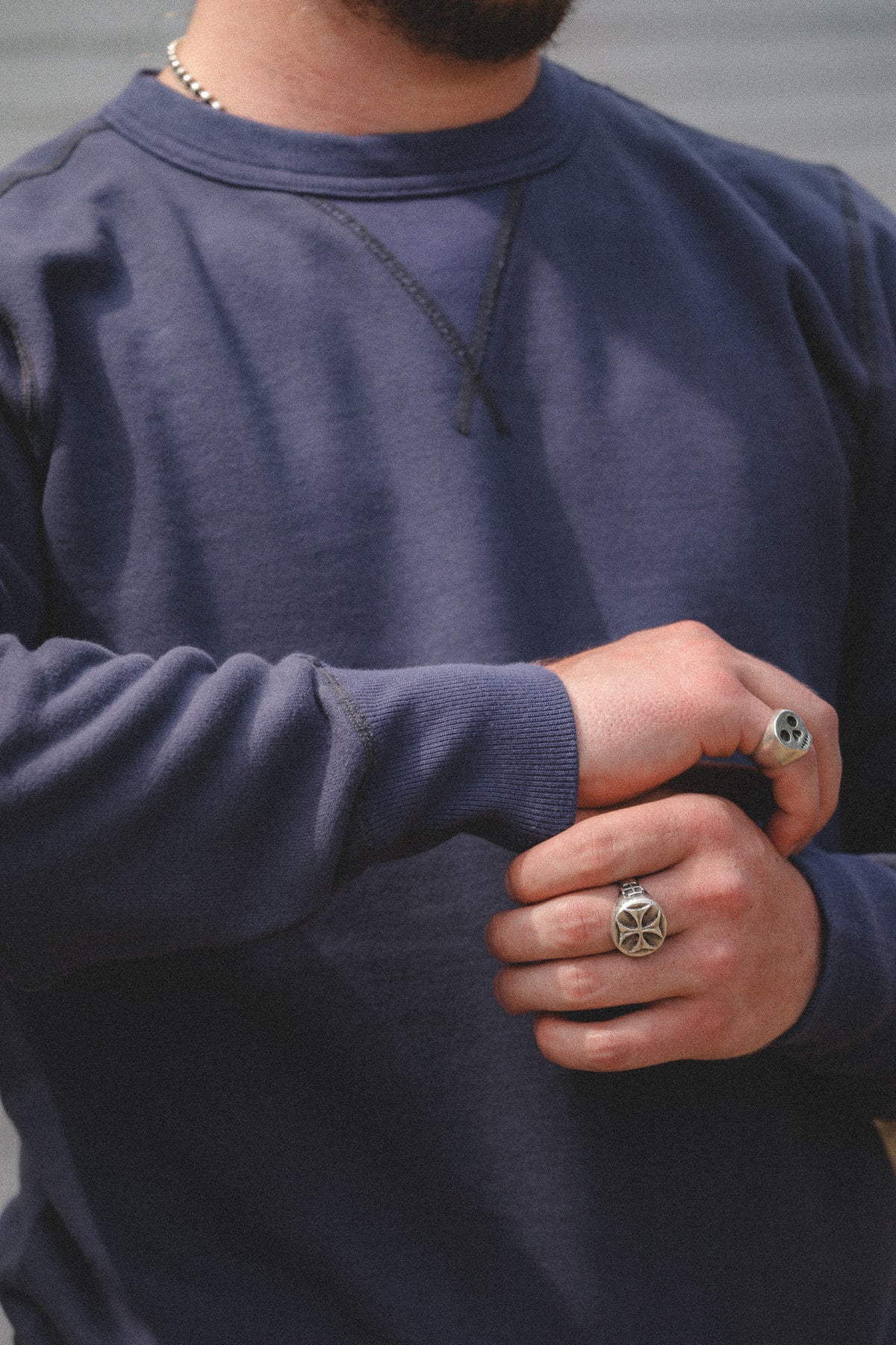 A close-up of a person wearing a navy blue crewneck sweatshirt, showing the chest area with a black chain necklace and silver rings on the fingers.