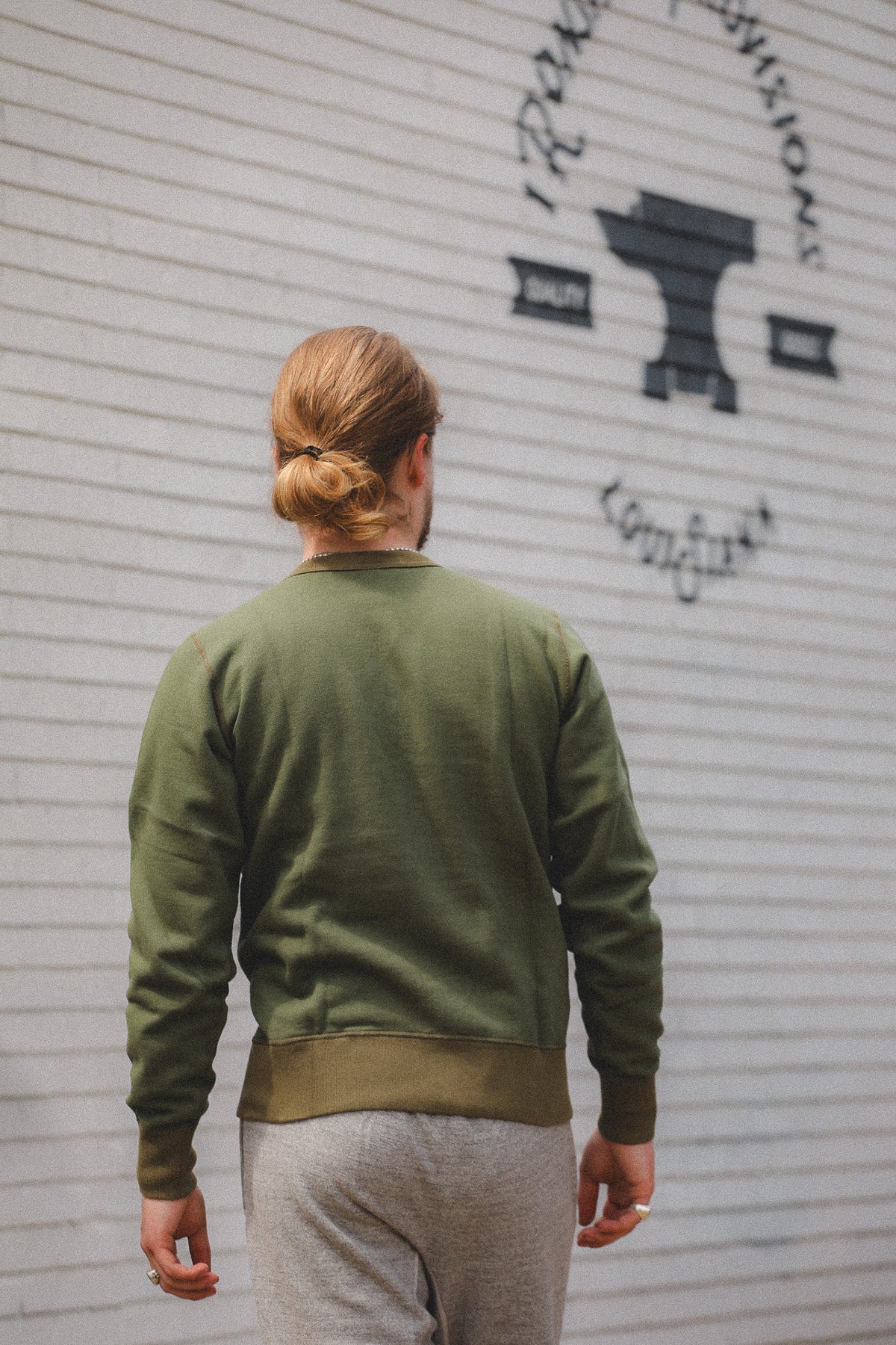 A man viewed from behind wearing an olive green crewneck sweatshirt and gray sweatpants, standing in front of a white brick wall with a black anvil logo.