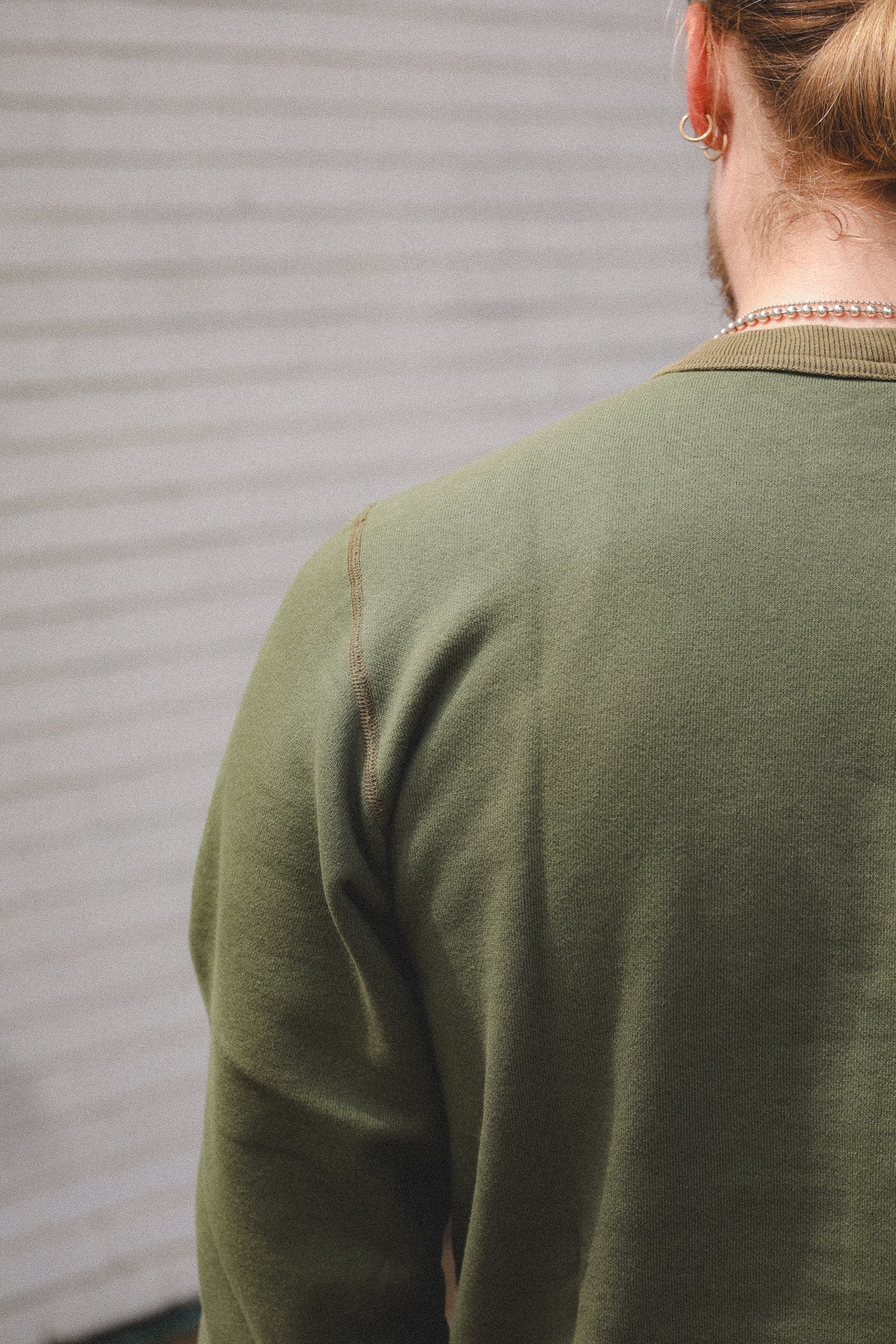Close-up back view of a person wearing an olive green knit sweater against a light gray corrugated metal wall.