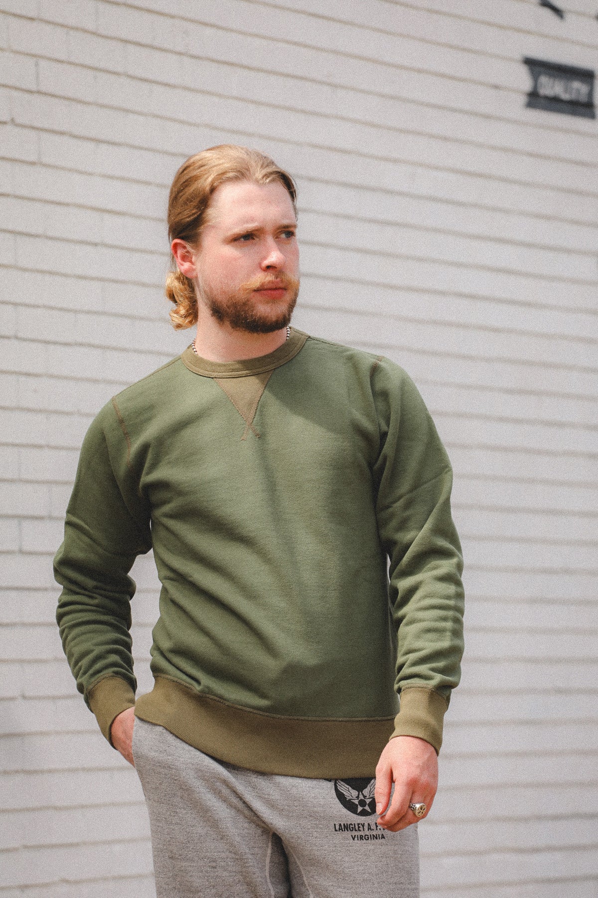 A man with blonde hair tied back and a beard wearing an olive green crewneck sweatshirt and gray sweatpants, standing against a white brick wall.