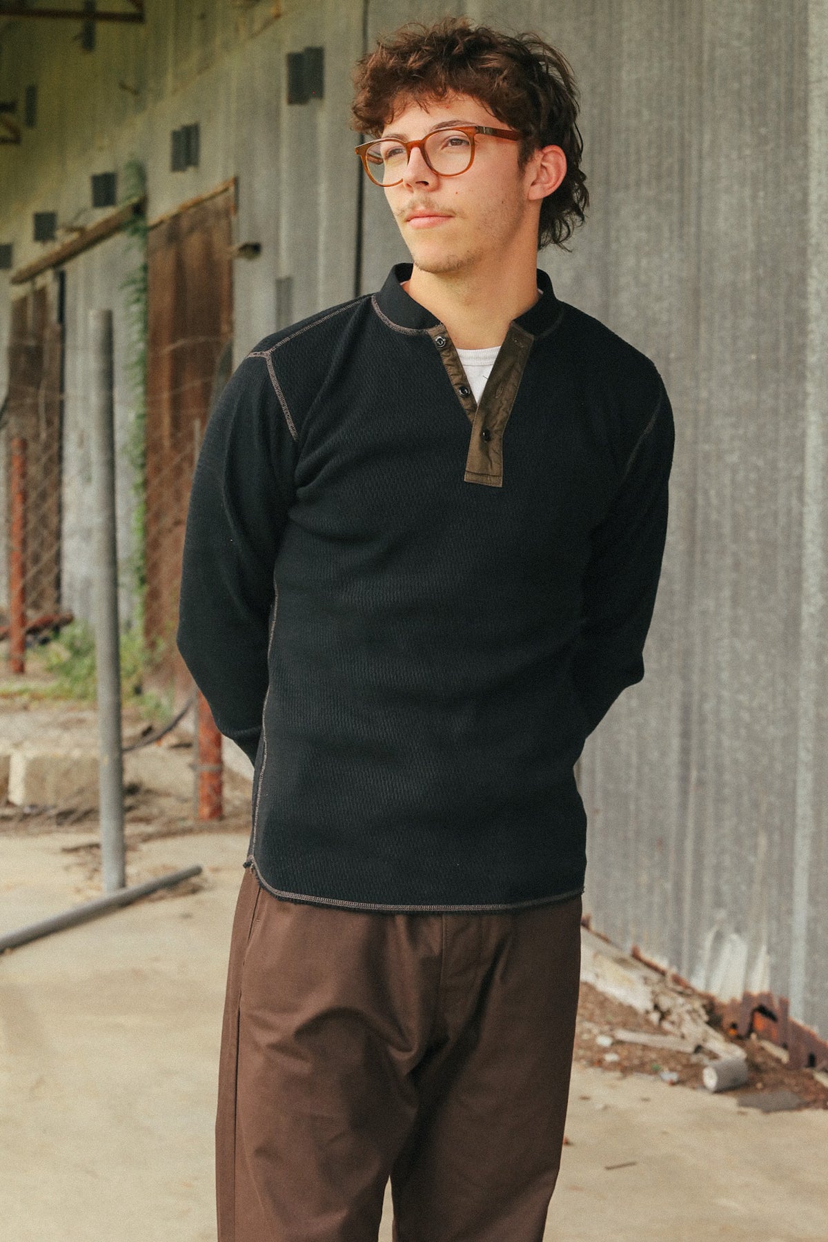 A man wearing a dark charcoal long-sleeve quarter-zip pullover with brown contrast stitching and a brown collar, paired with brown pants, standing in front of a corrugated metal wall.
