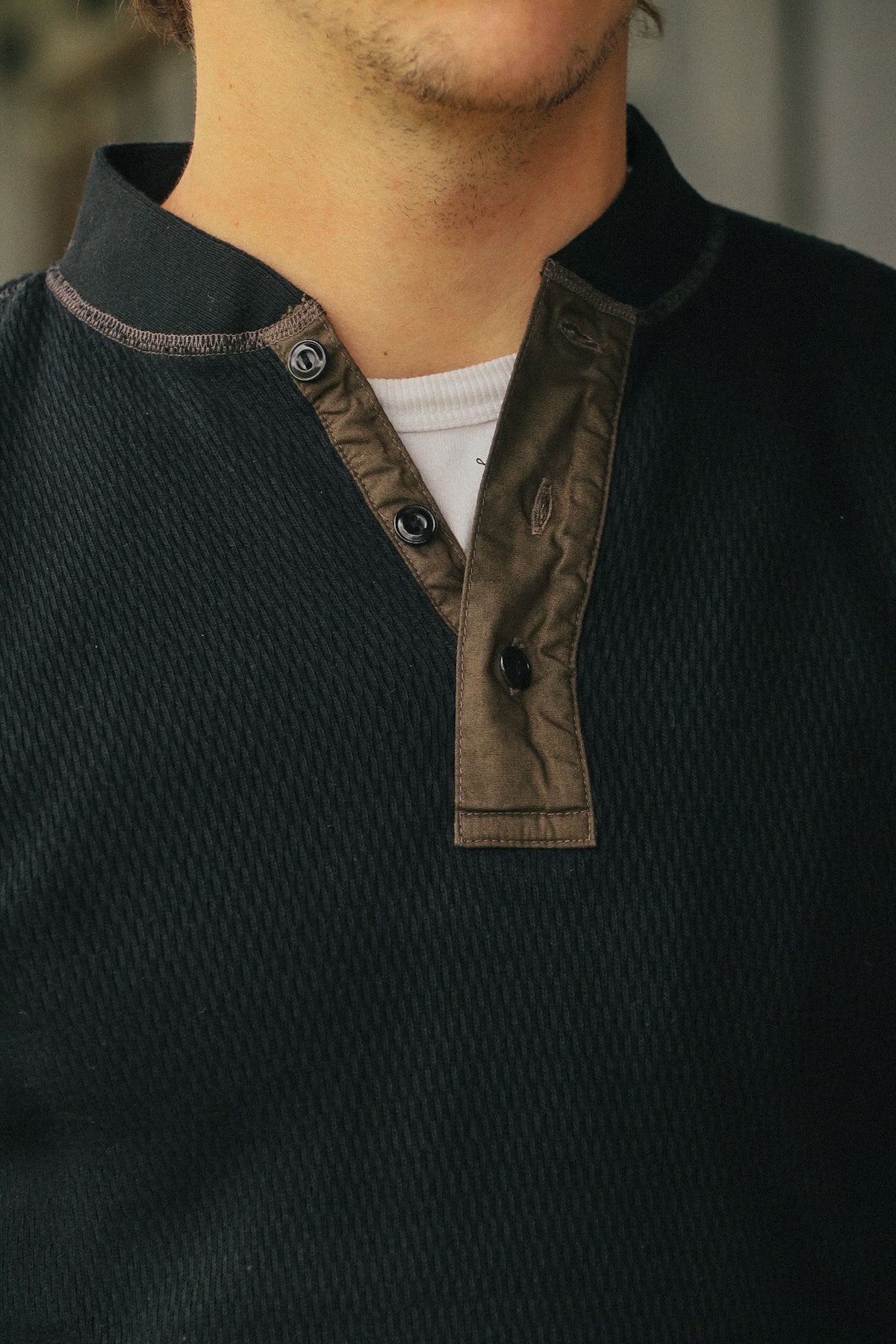 Close-up of a person wearing a dark charcoal knit Henley shirt with a brown contrast placket and collar trim, layered over a white undershirt.