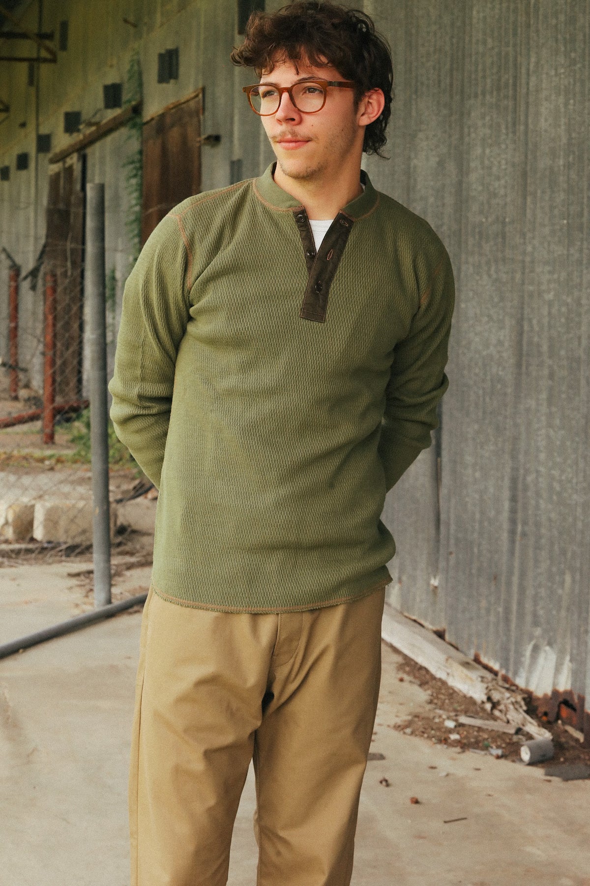 A man wearing a textured olive green long-sleeve henley sweater and tan pants, standing outdoors against a corrugated metal wall.