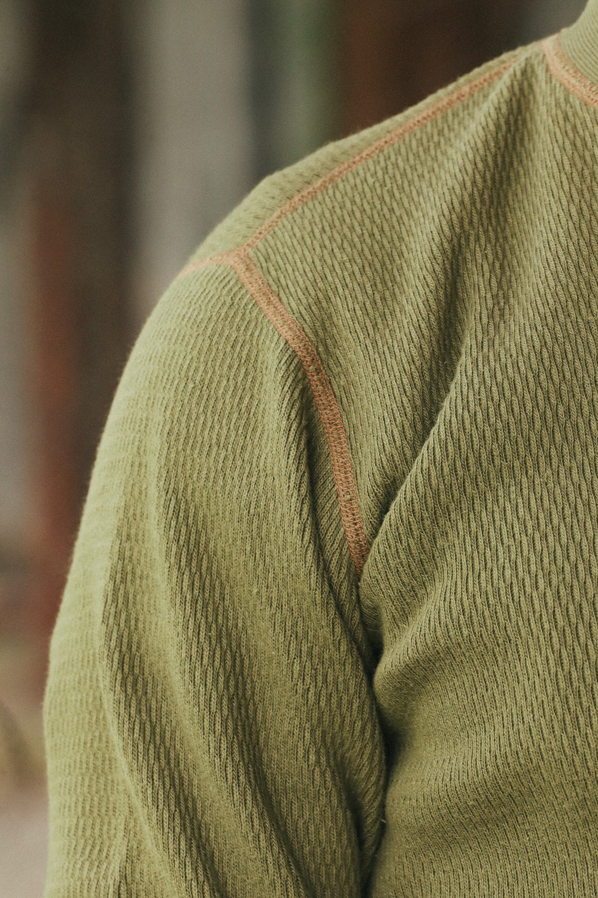 Close-up of a textured olive green knit garment, showing the shoulder and upper arm with contrasting peach-pink seam stitching against a blurred outdoor background.