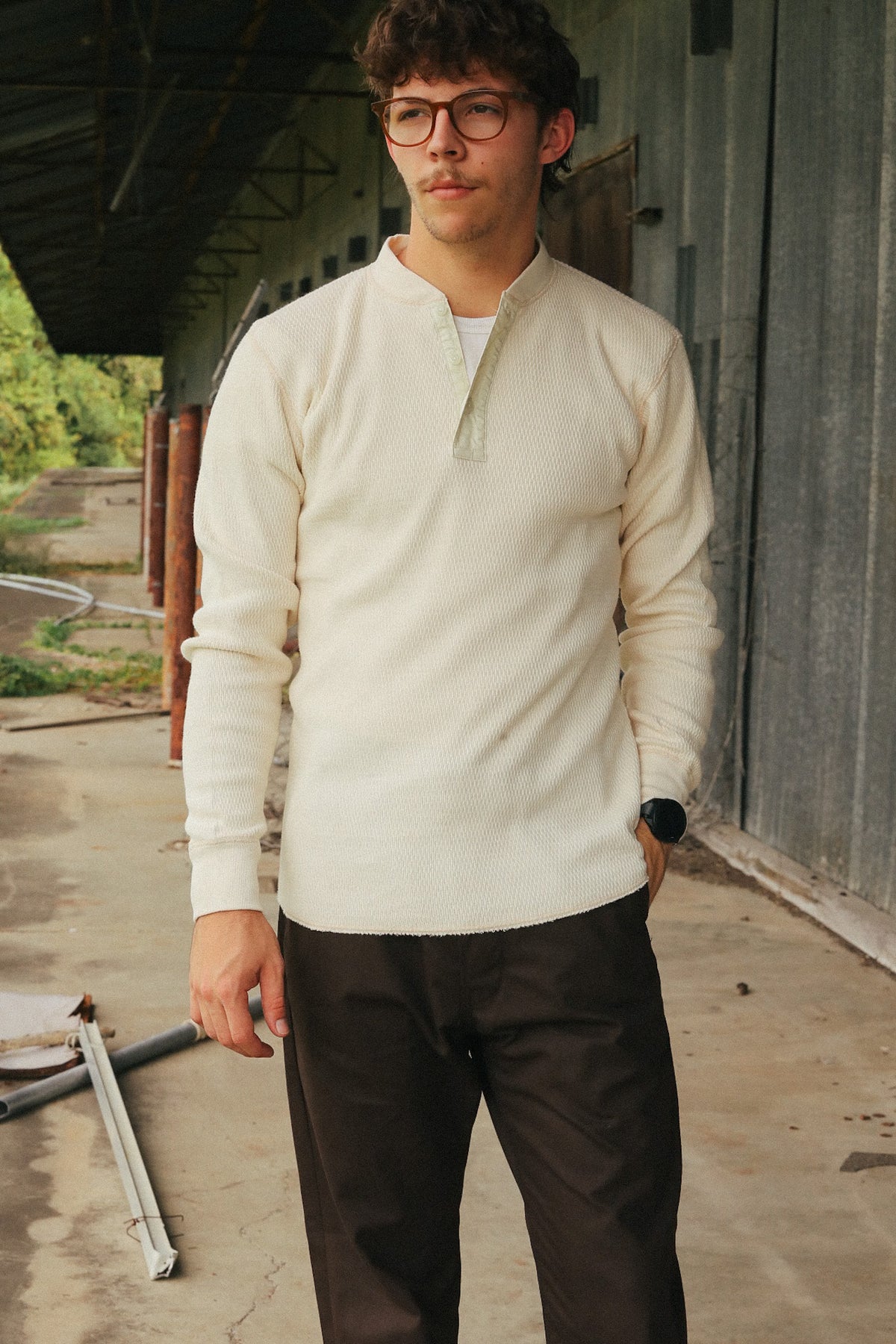 A man wearing a cream-colored long-sleeve quarter-zip henley sweater and dark brown pants, standing in front of a rustic metal building.