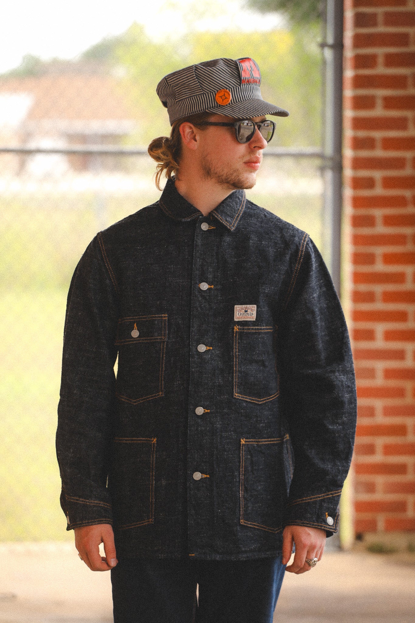 Man wearing a dark denim jacket and cap, standing outdoors near a brick building.