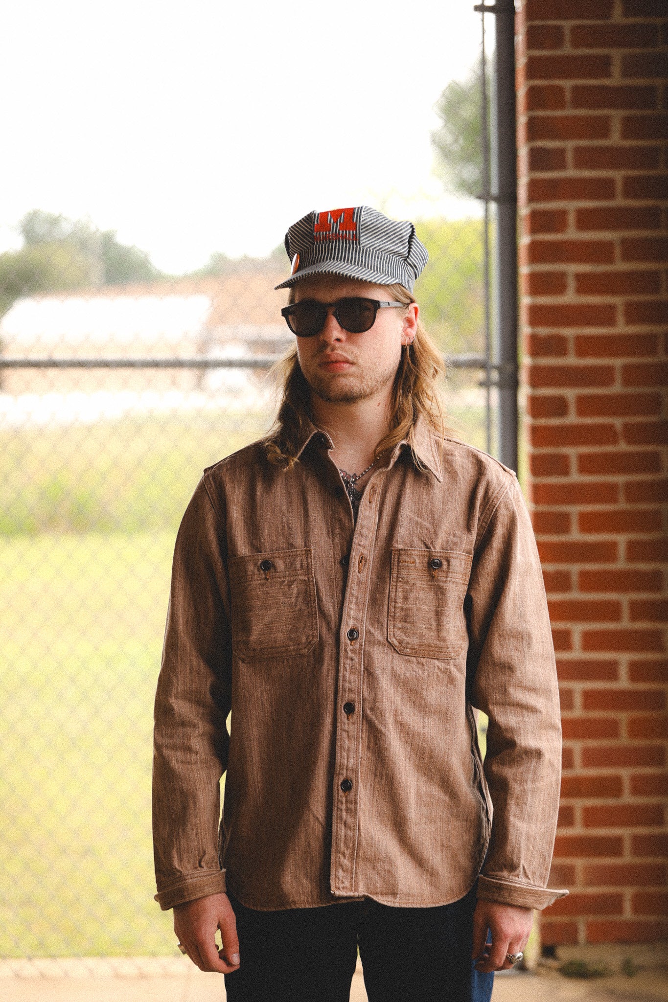 Man wearing a brown jacket, sunglasses, and a cap standing in front of a brick wall.