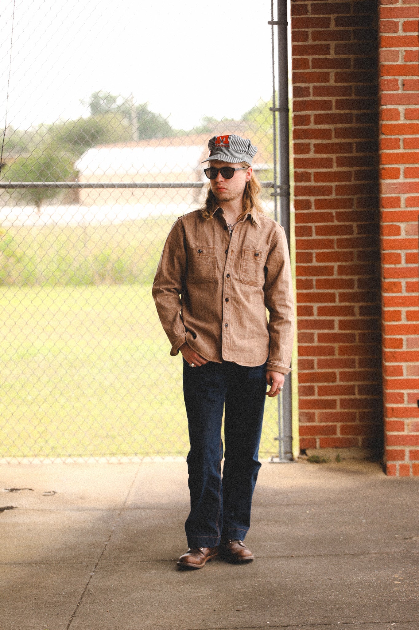 Person wearing a brown shirt and blue jeans standing against a brick wall.