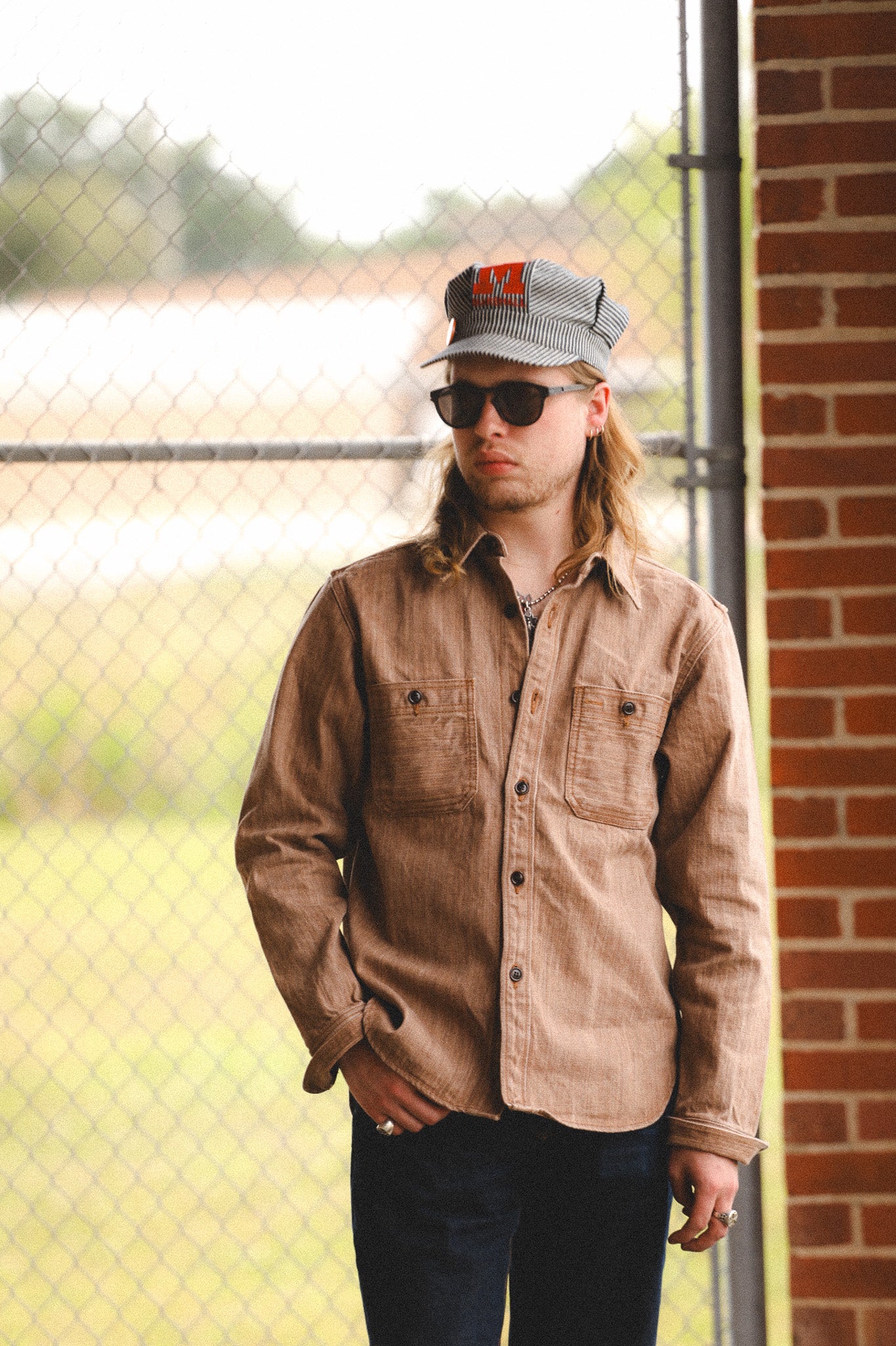 Person wearing a brown jacket and sunglasses standing near a brick wall.