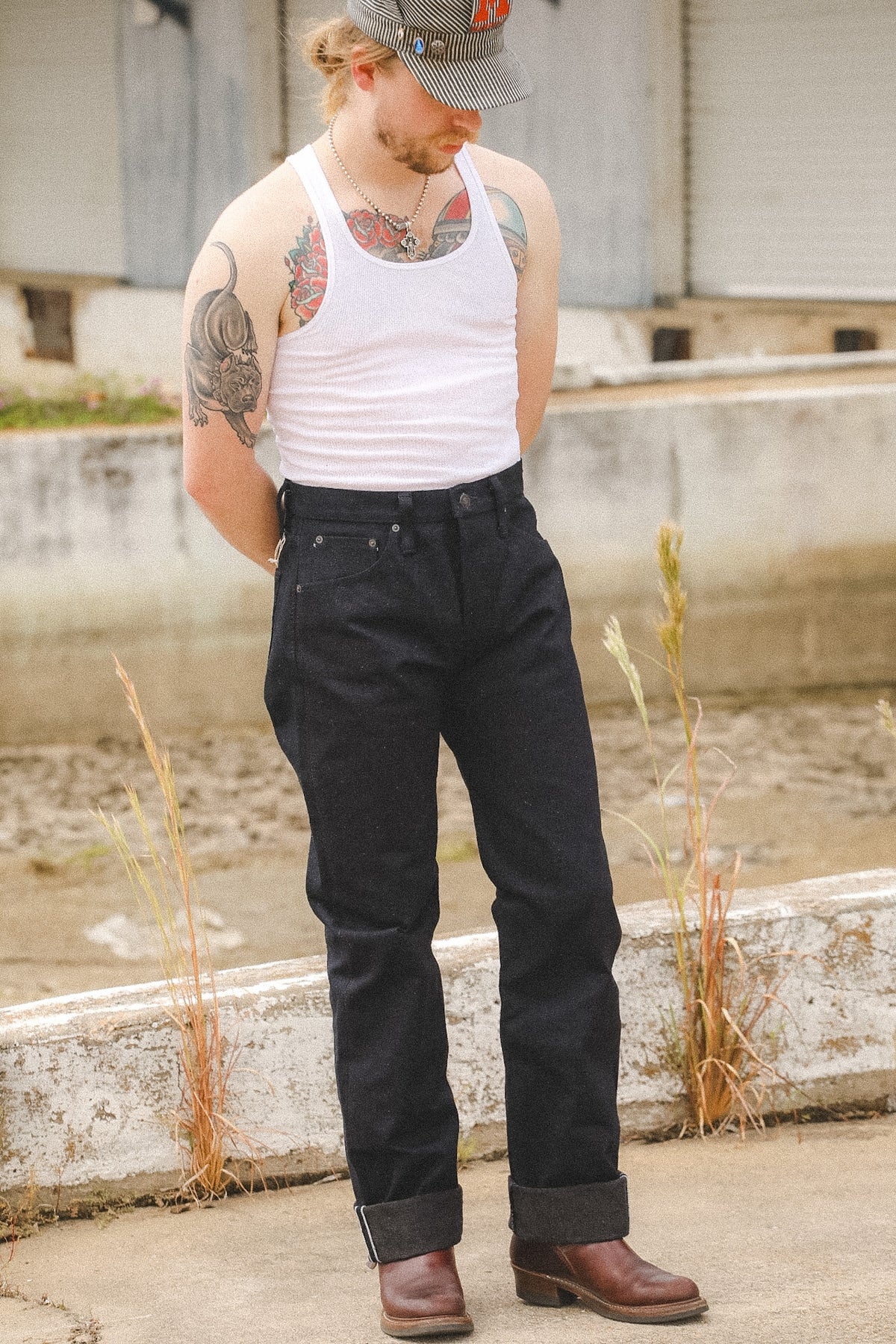 A man wearing dark wash denim jeans, a white tank top, and brown boots, standing outdoors in front of a concrete wall.