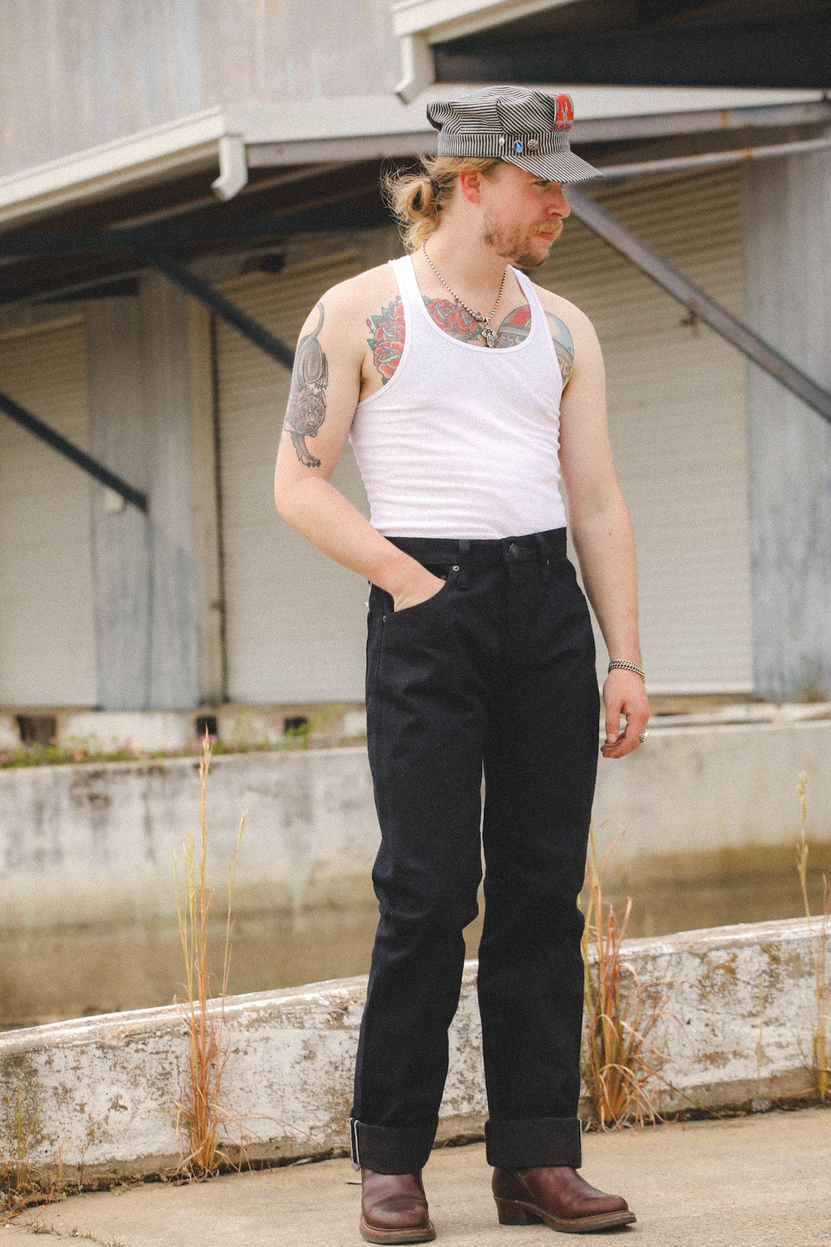 A man wearing a white ribbed tank top, black wide-leg jeans, brown boots, and a gray cap stands outdoors in front of a concrete wall and industrial building.