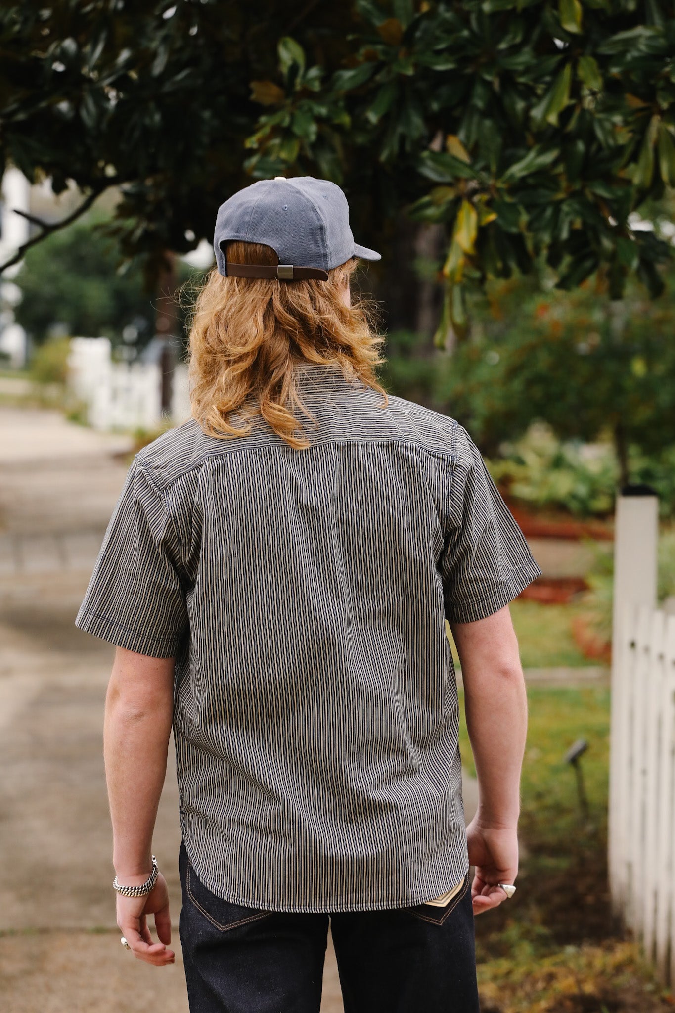 Person wearing a gray checkered shirt and cap walking outdoors with greenery in the background