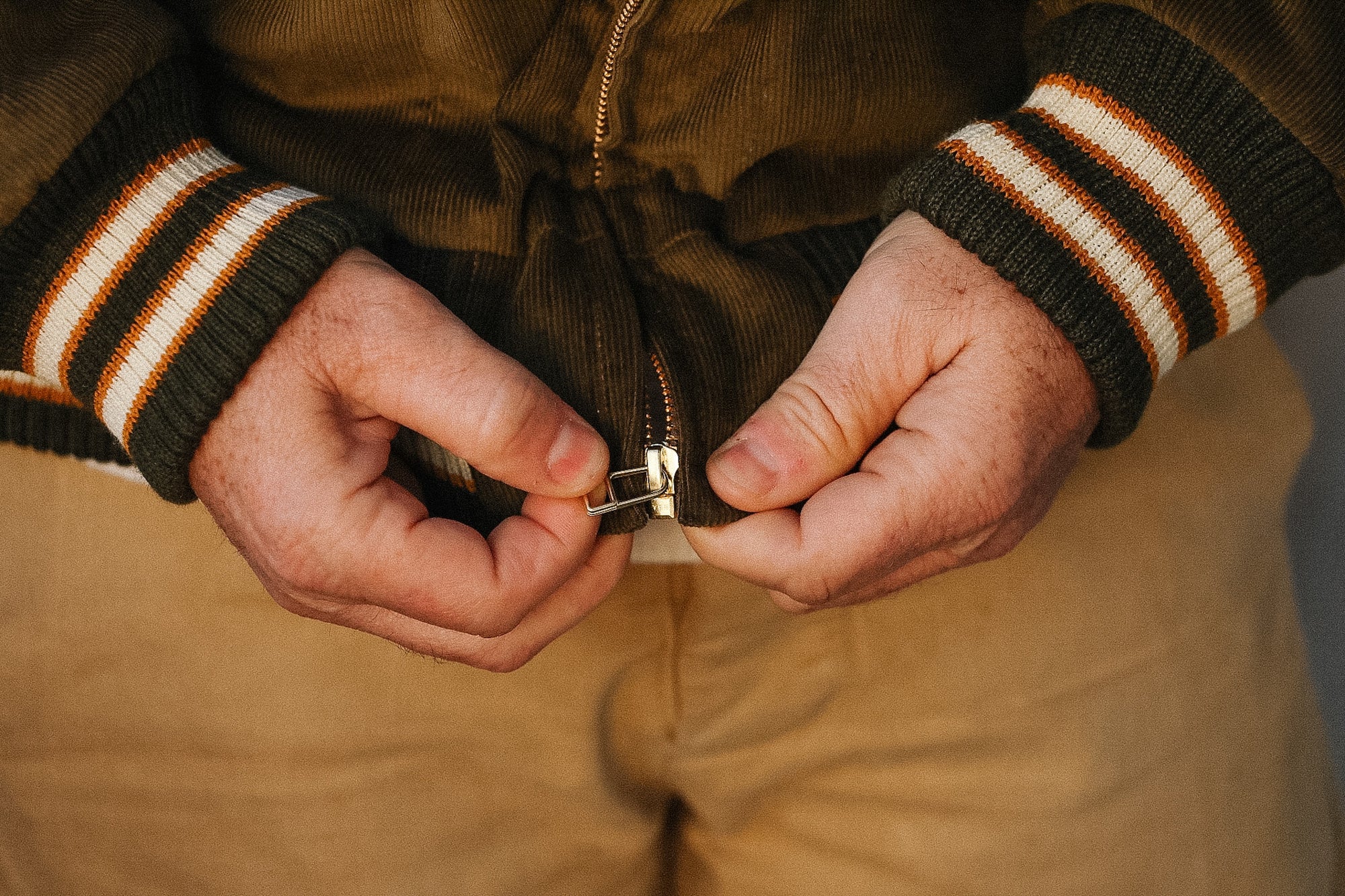 Person wearing a brown jacket with striped cuffs adjusting a zipper.