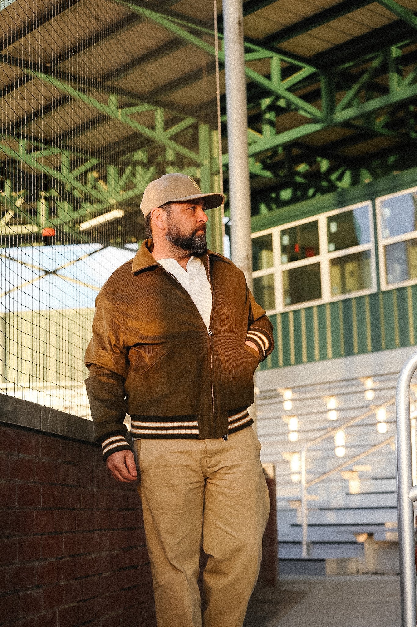 Man wearing a brown jacket with striped sleeves standing outdoors under a green metal structure.
