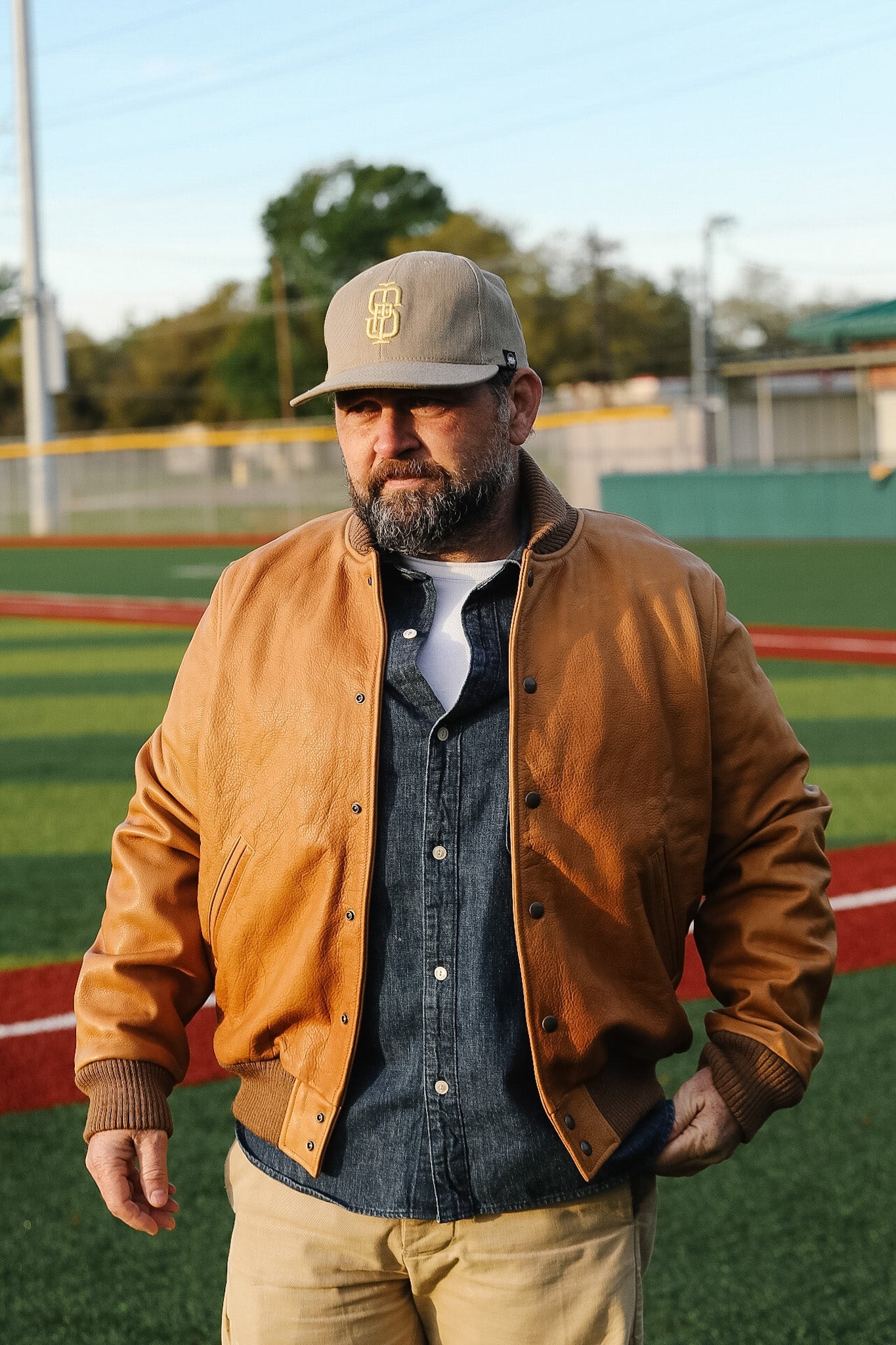 Man wearing a brown leather jacket and cap on a sports field