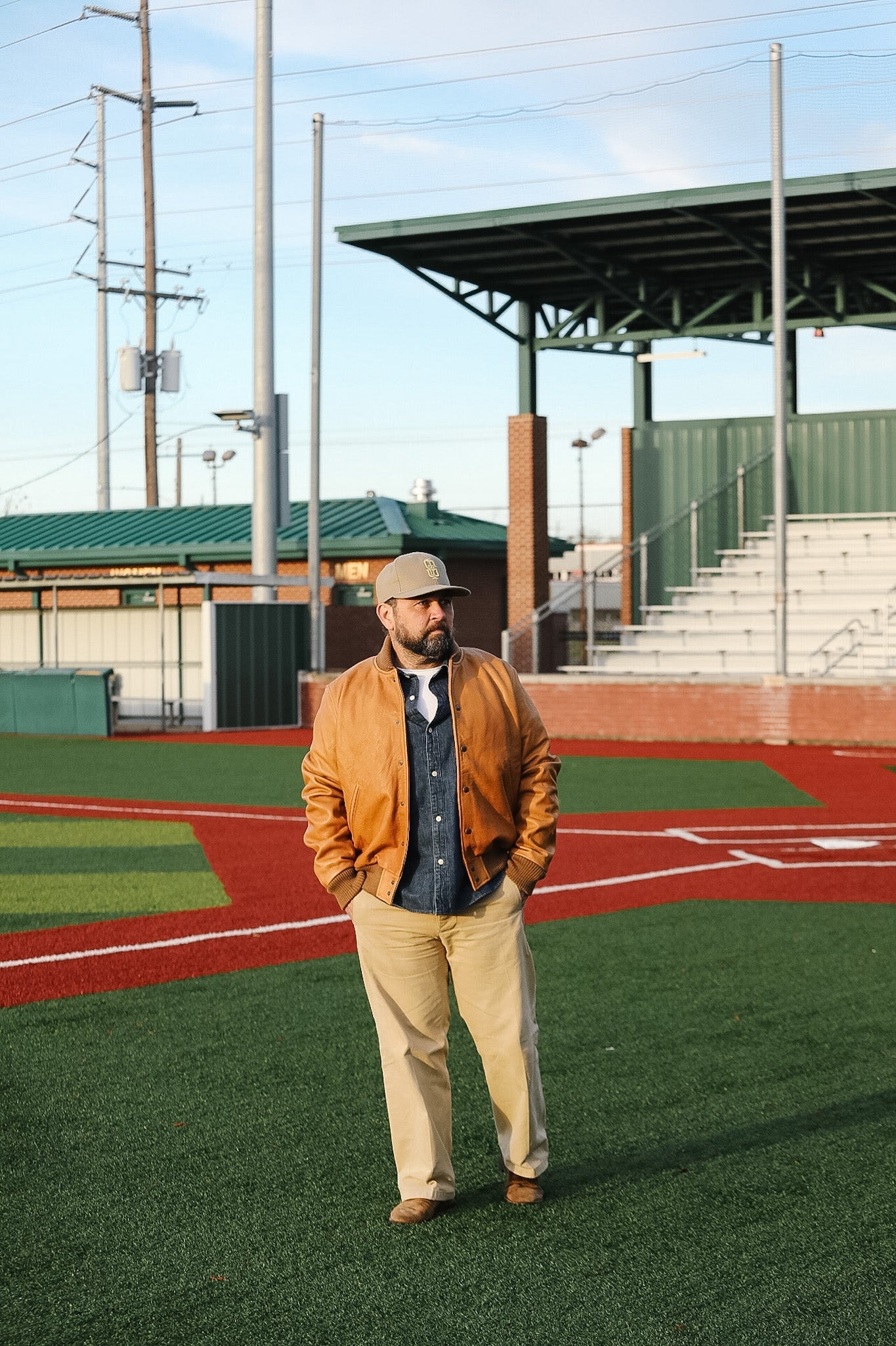 Man standing on a baseball field wearing a brown jacket and beige pants.