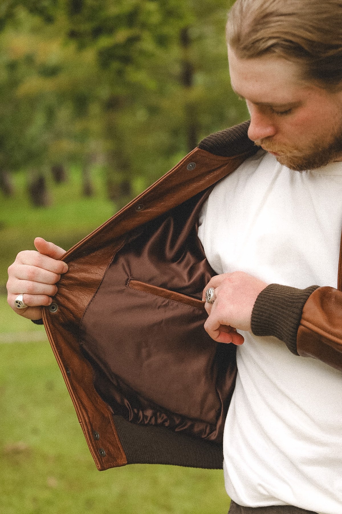 A man outdoors wearing a brown leather jacket and opening it to show the brown satin-lined interior pocket.