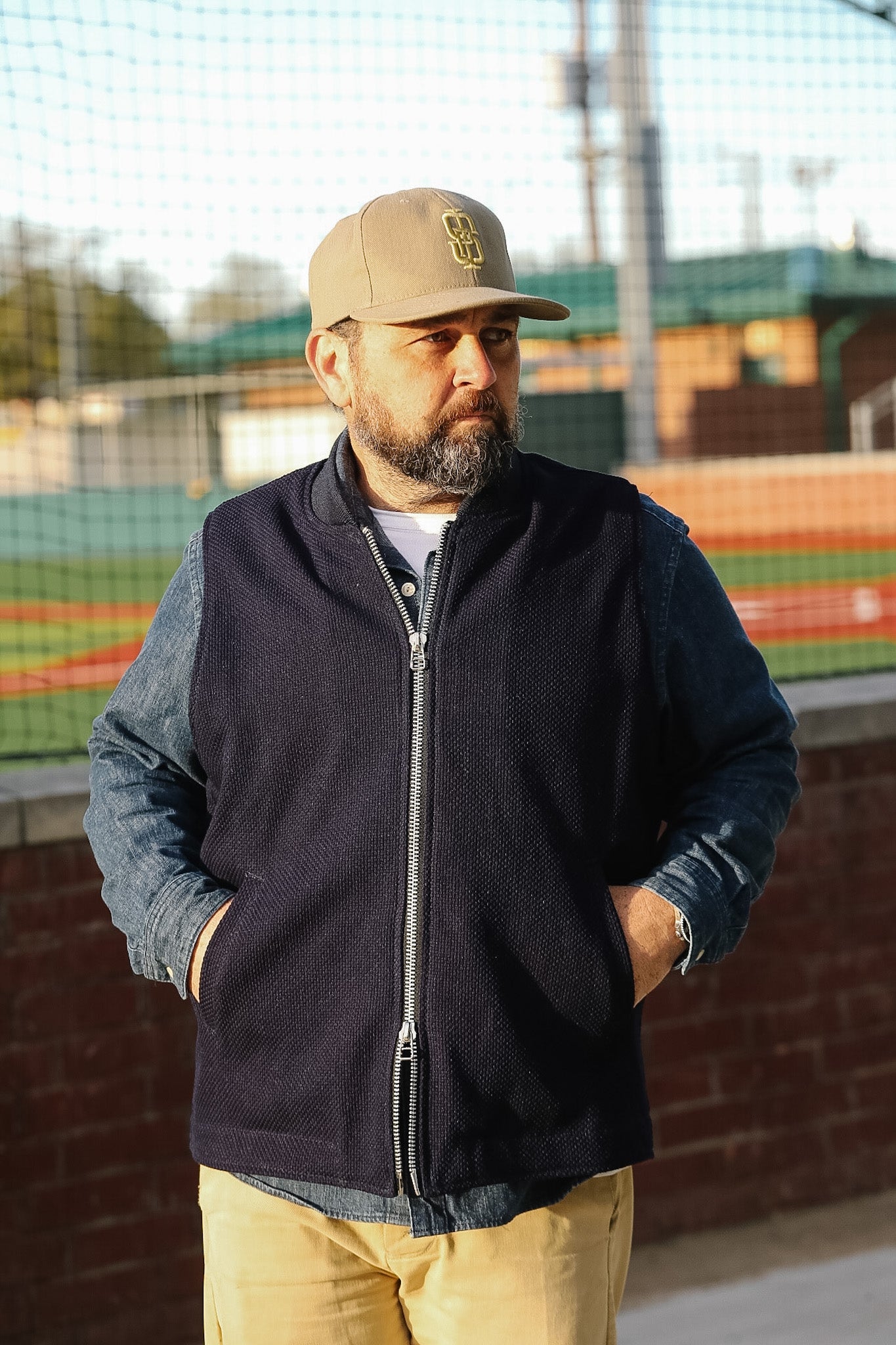 Man wearing a black jacket and beige cap standing outdoors with a sports field in the background