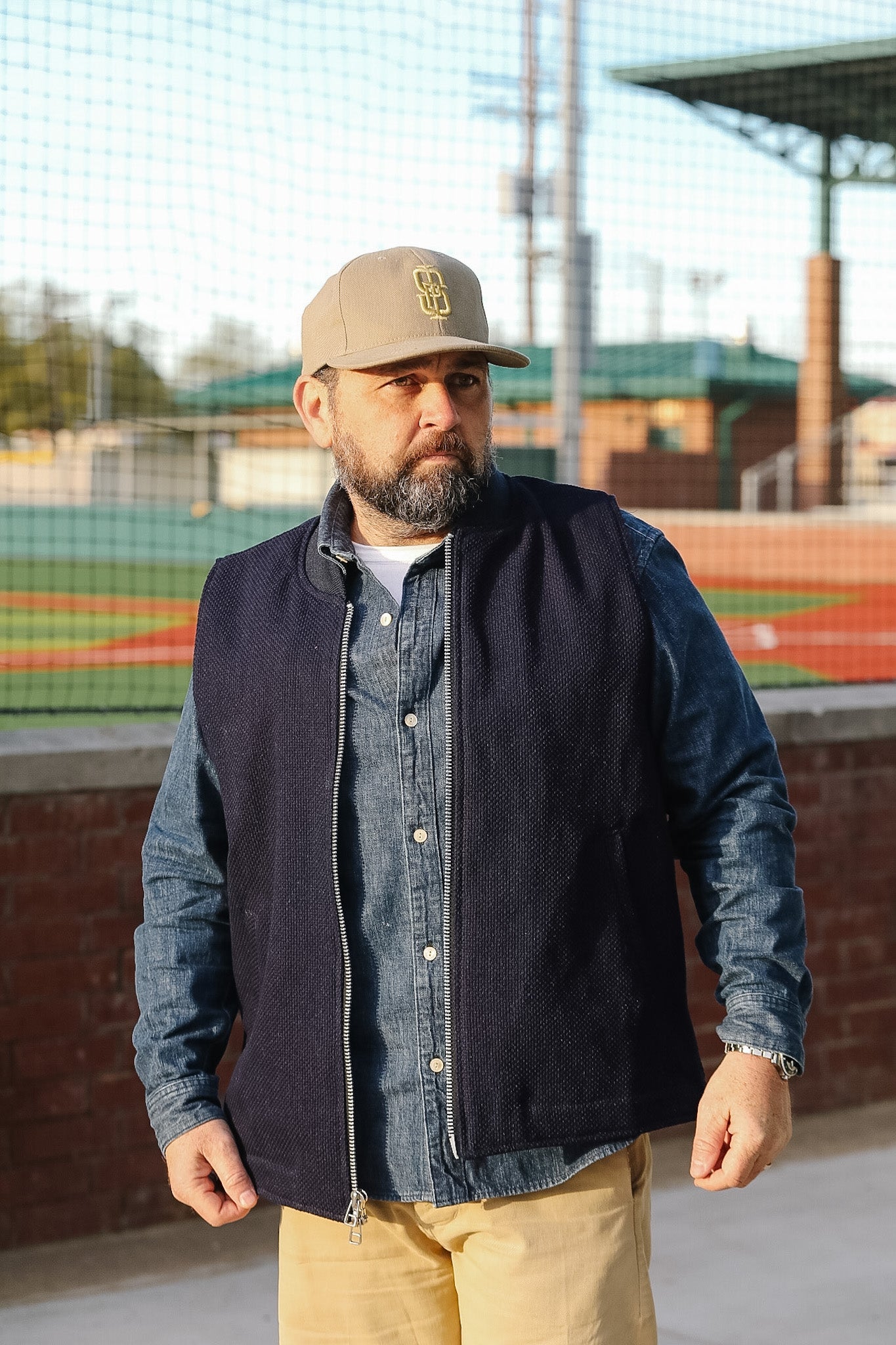 Man wearing a navy jacket, denim shirt, beige pants, and a beige cap outdoors.