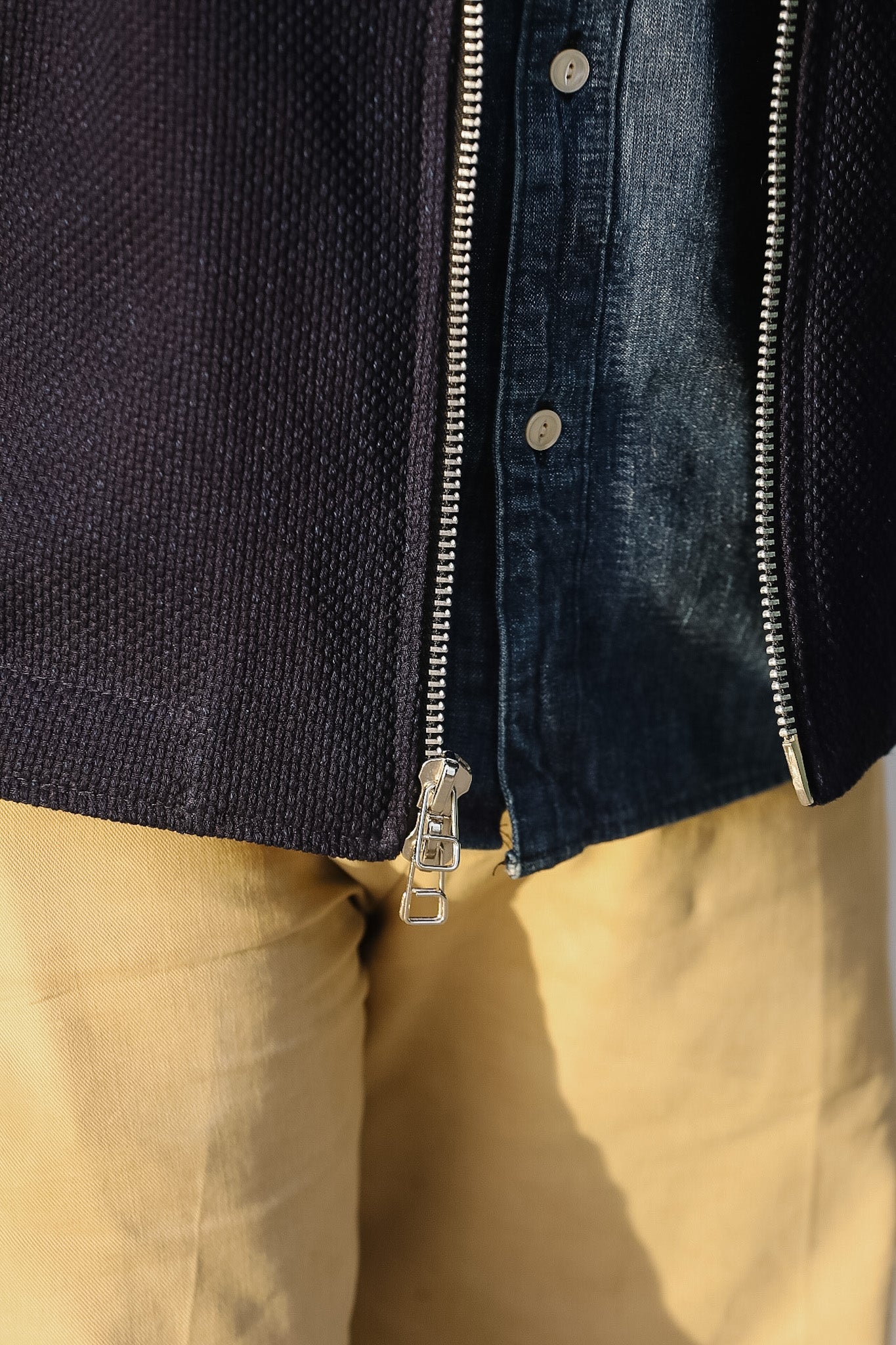 Close-up of a person wearing a dark jacket with a zipper, beige pants, and a blue shirt.
