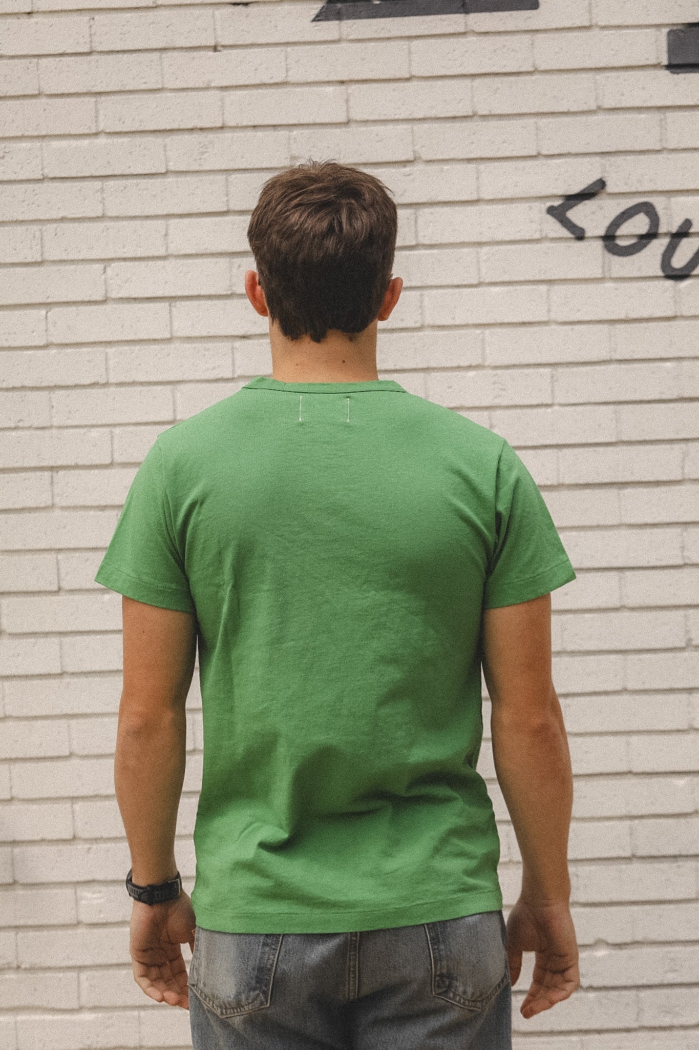 Person wearing a green t-shirt standing against a light brick wall with graffiti.