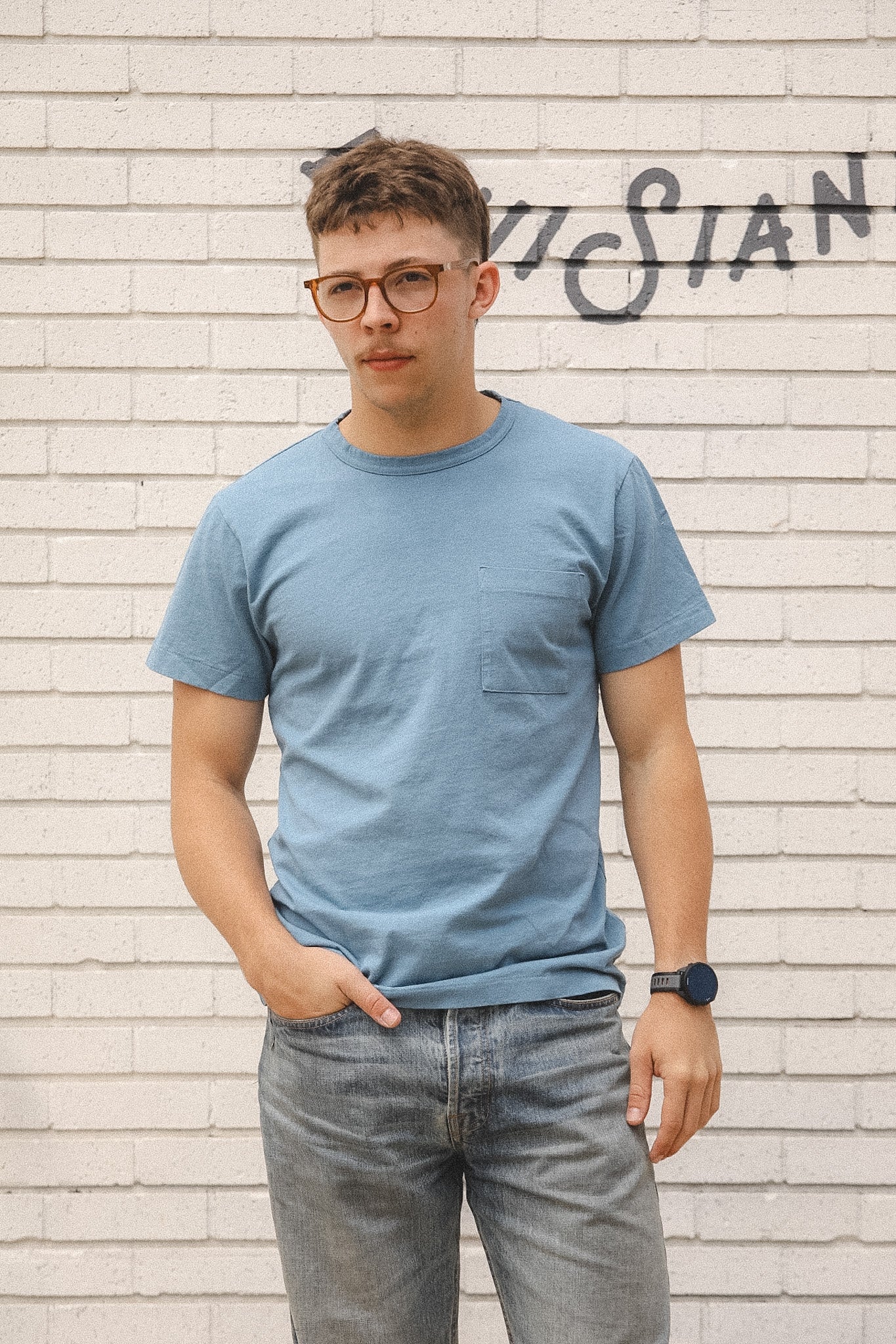 Man wearing a blue t-shirt and glasses standing against a textured wall.