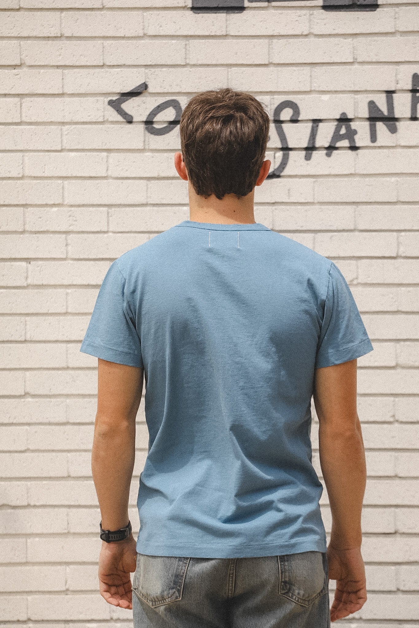 Person wearing a blue t-shirt standing against a light brick wall with graffiti.