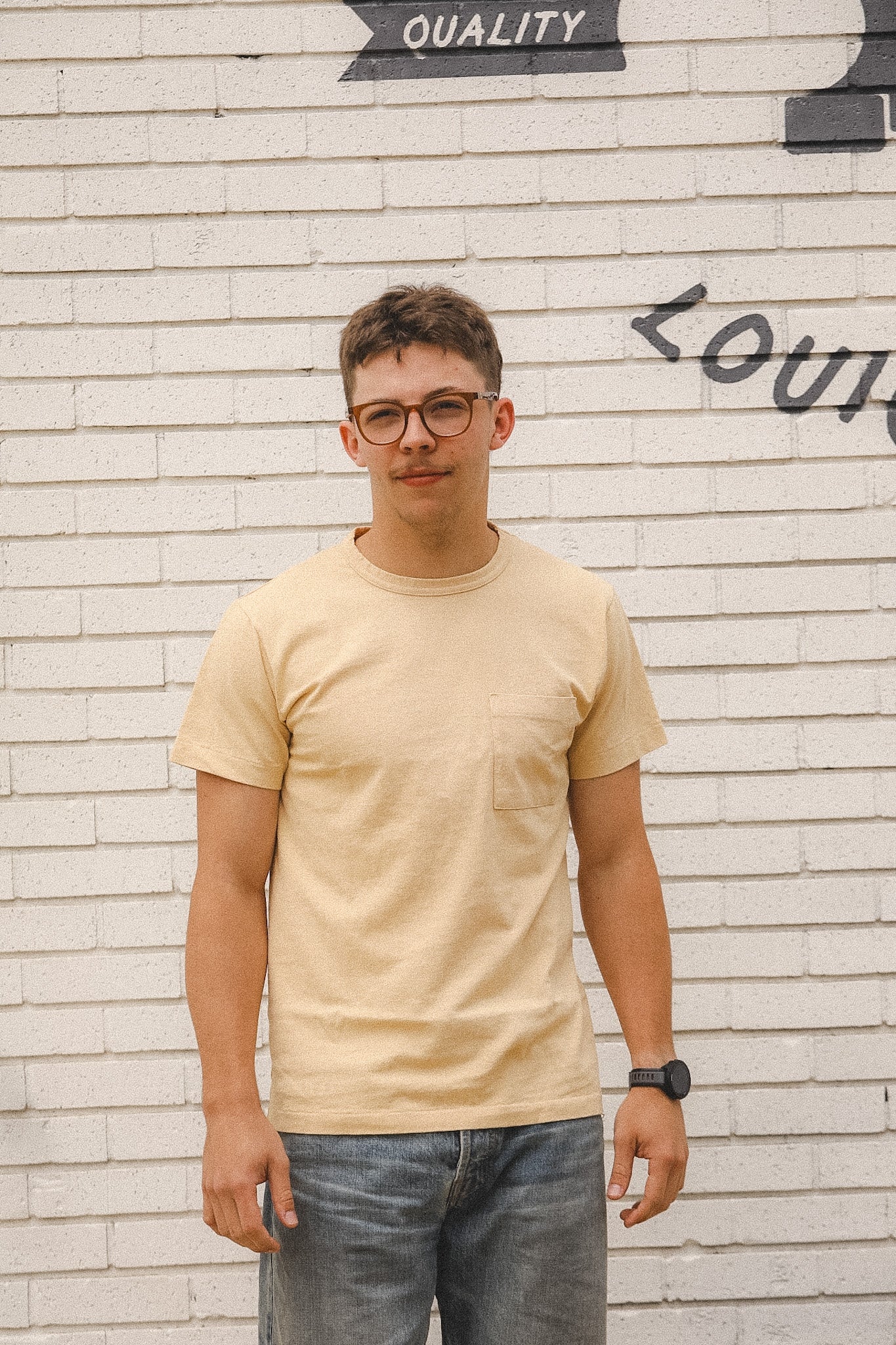 Person wearing a beige t-shirt and glasses standing in front of a textured wall.