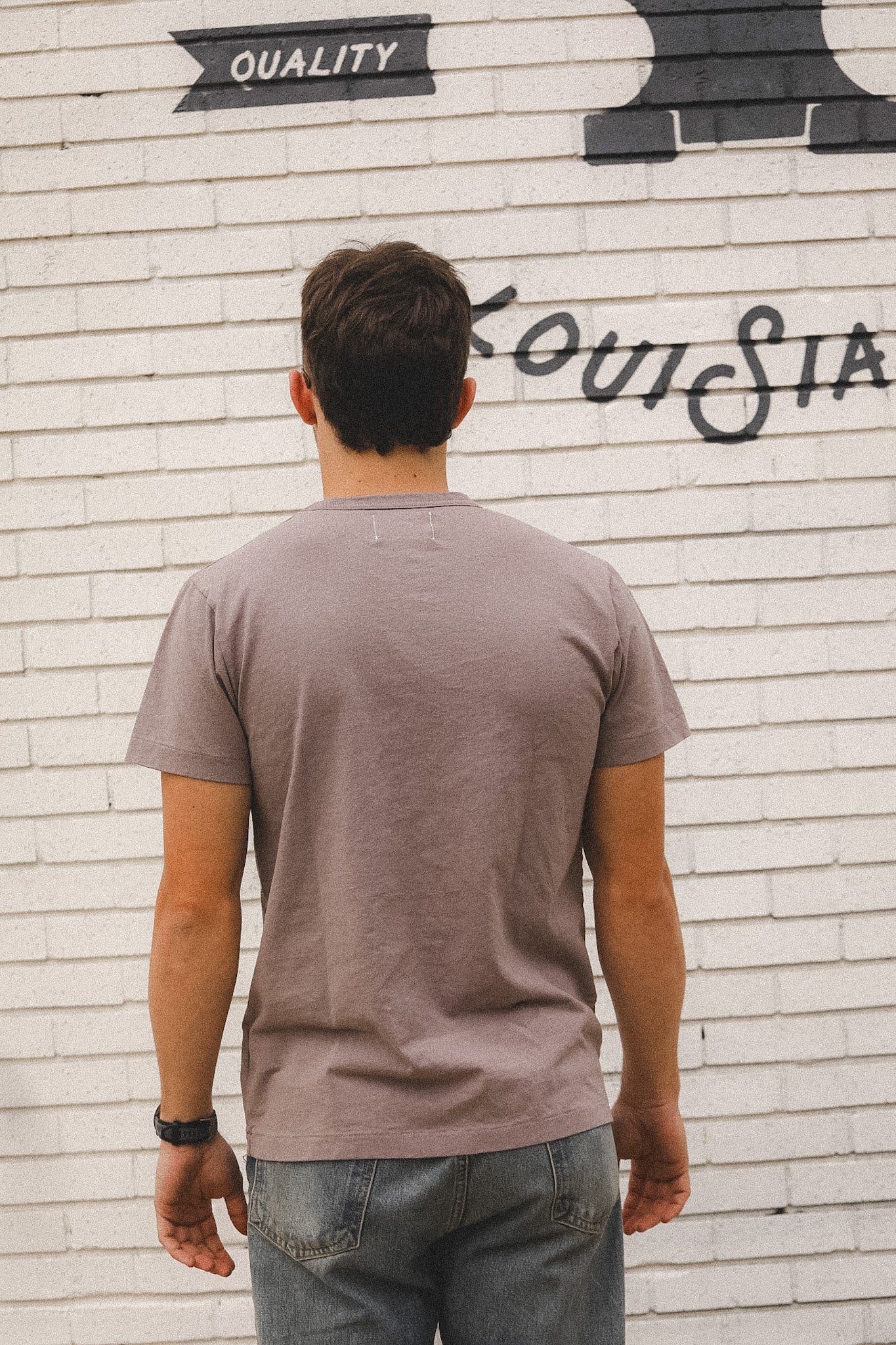 Man wearing a gray t-shirt standing in front of a white brick wall with graffiti.