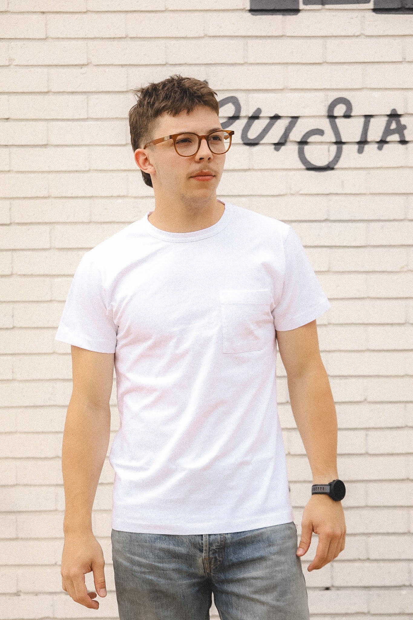 Man wearing a white t-shirt and glasses standing against a textured wall with graffiti.